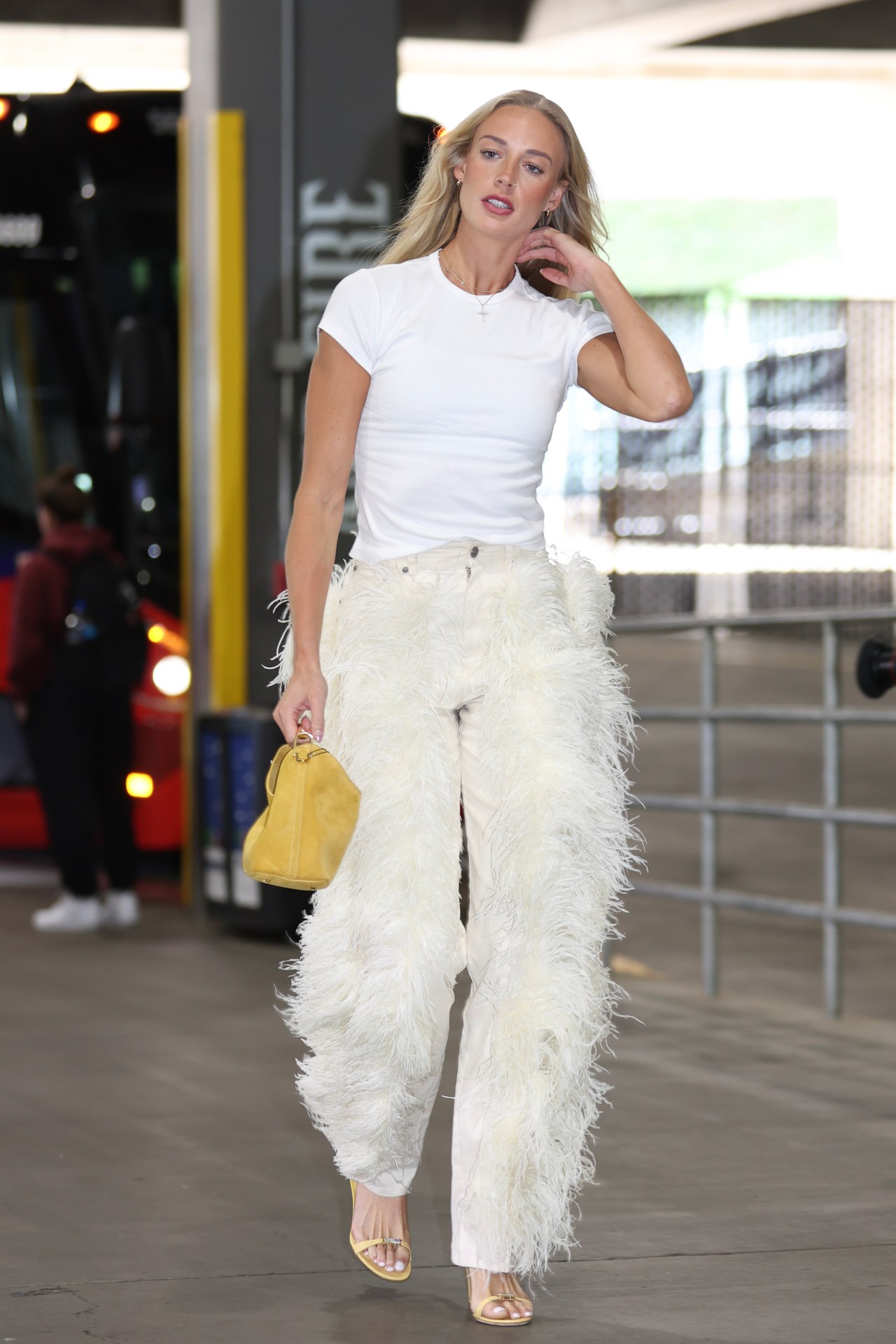 Sophie Cunningham wearing white feather-trimmed pants and a white t-shirt, carrying a yellow handbag, walking into Moda Center before a game.