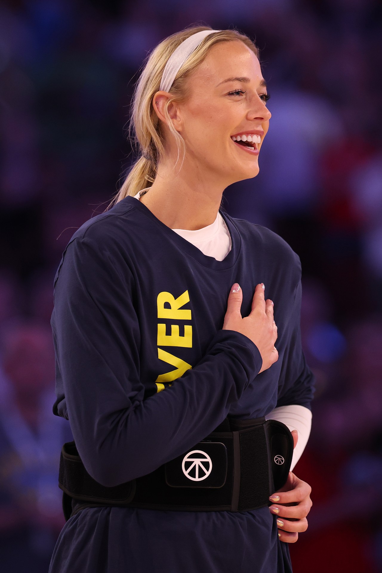 Blonde woman in navy Indiana Fever warmup shirt holds her hand over her heart and smiles during the national anthem.
