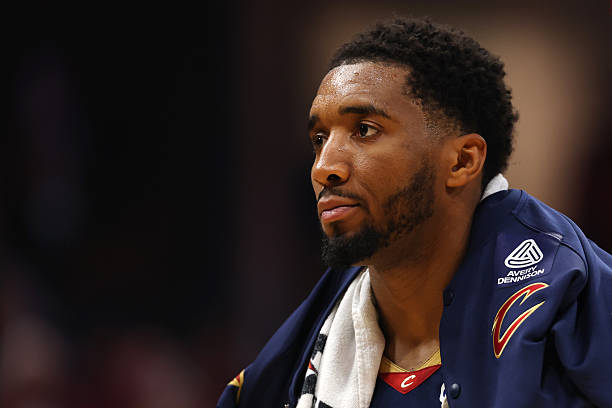 Donovan Mitchell in a navy Cavaliers jacket with a towel around his neck, gazing to the side during an NBA game.