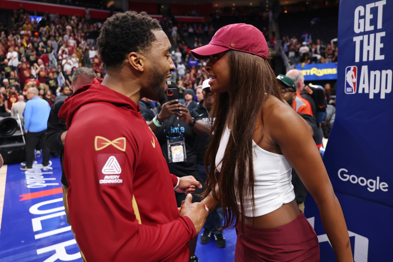 A man in a red Cavaliers hoodie and a woman in a white crop top and maroon cap hold hands and smile at each other on an NBA arena floor after a playoff game.