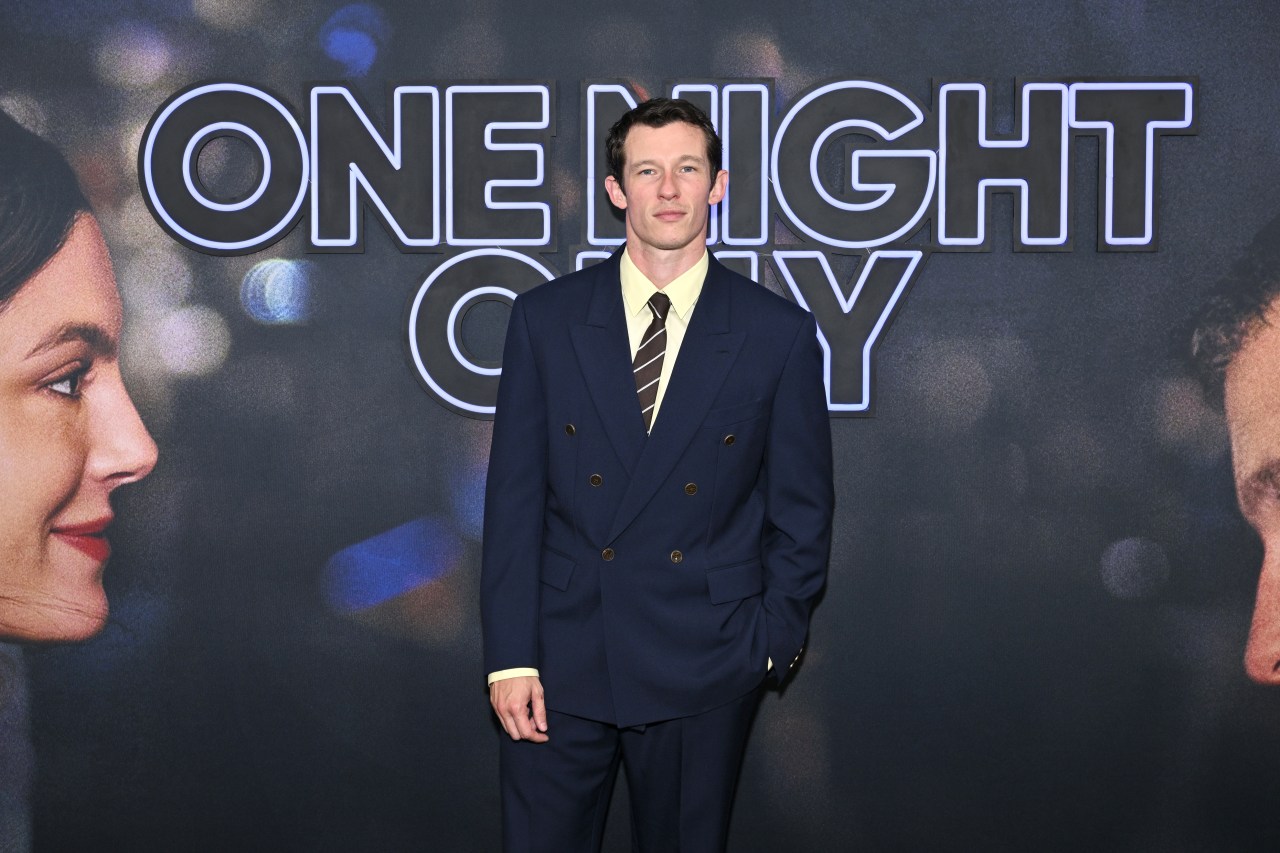 Callum Turner in a navy double-breasted suit, pale yellow shirt, and striped tie stands in front of a large One Night Only premiere backdrop.