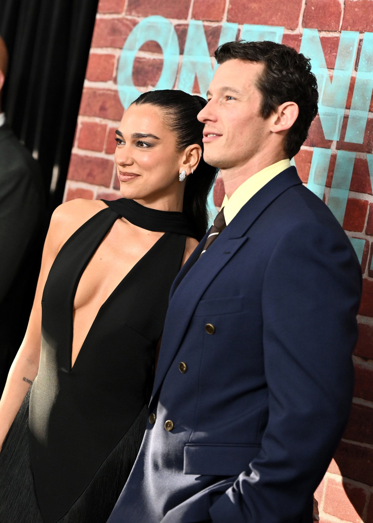 Dua Lipa in a black plunging halter gown and Callum Turner in a navy suit stand close together smiling in front of a brick wall at a film premiere.