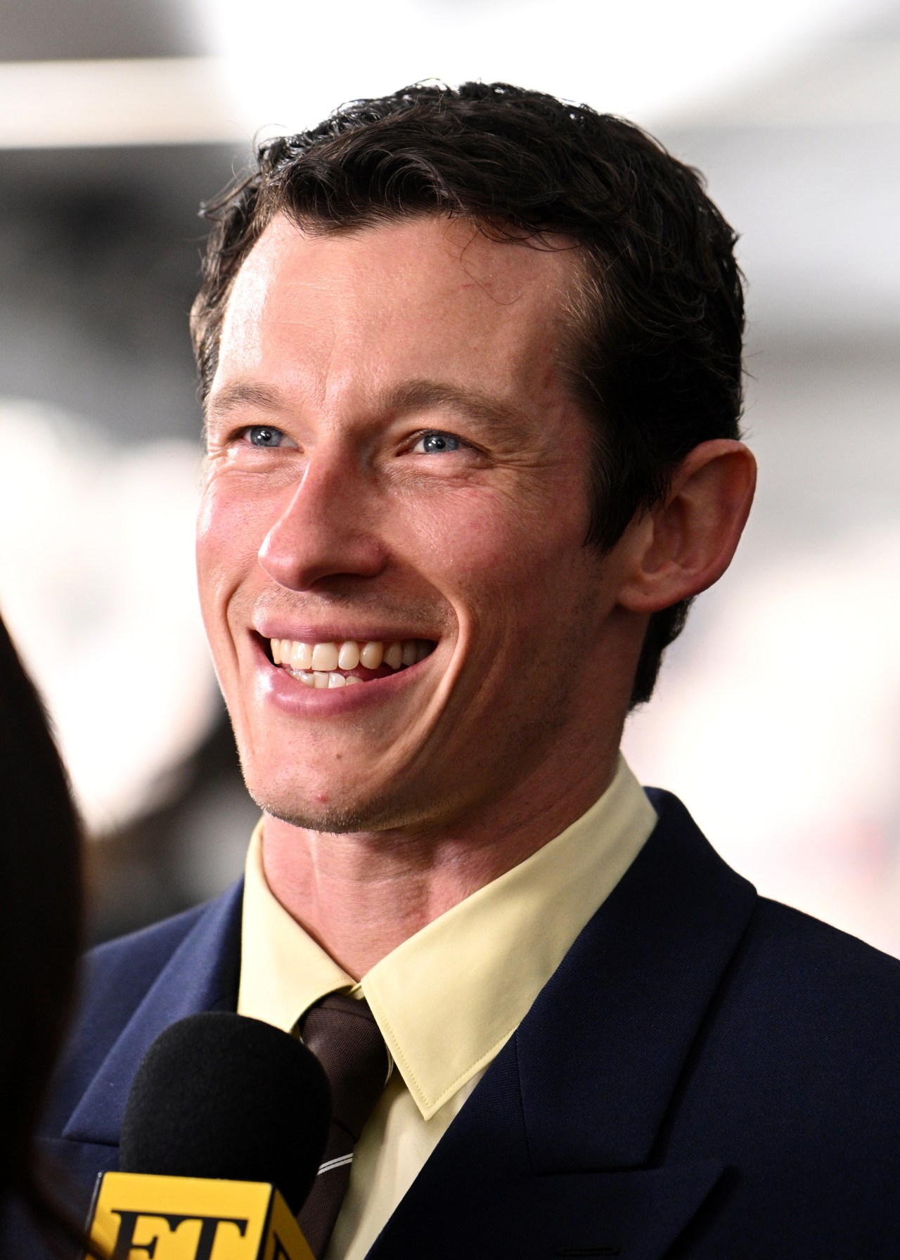 Callum Turner laughing while being interviewed, wearing a navy suit with a yellow shirt and brown tie.