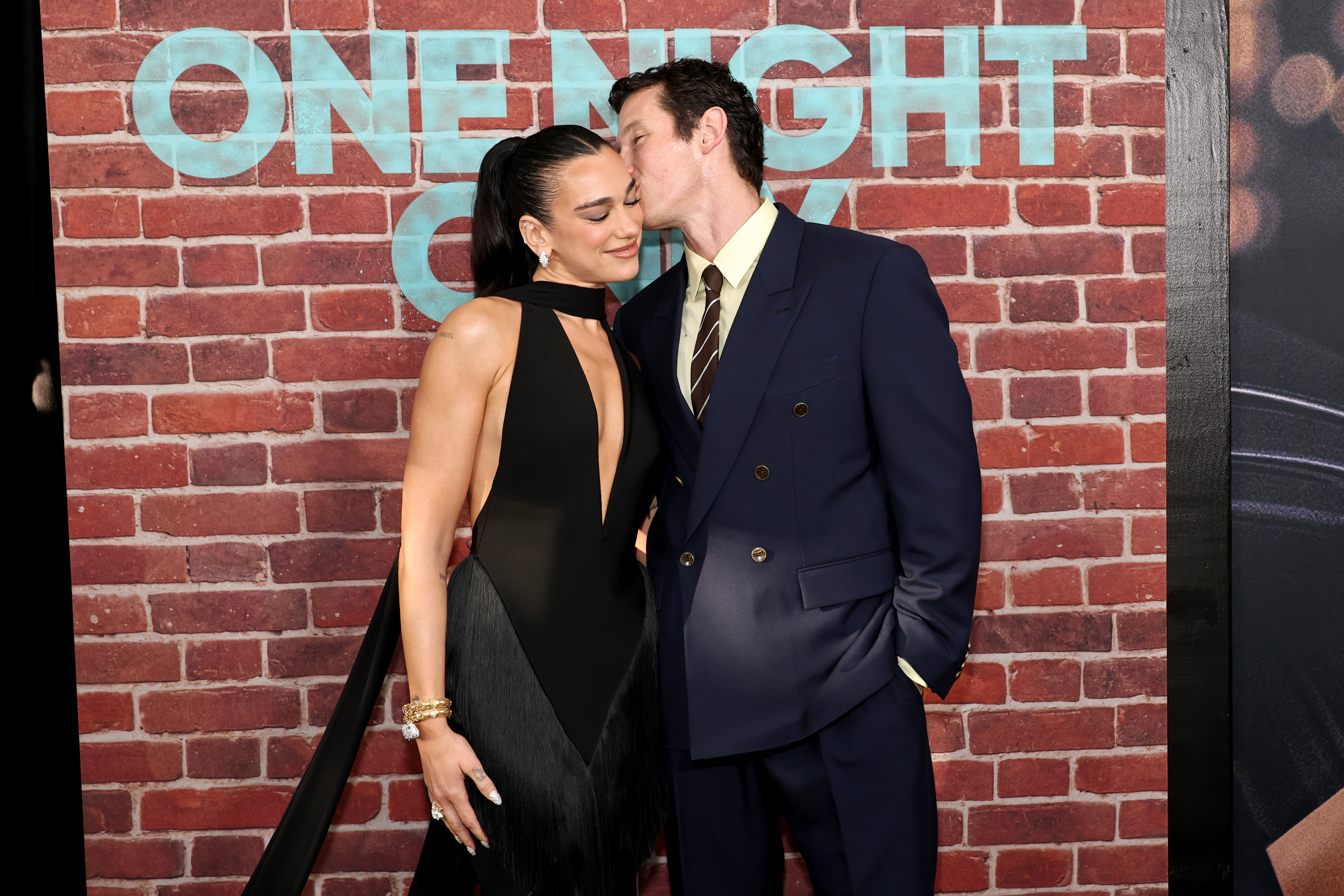 A man in a navy suit leans in to kiss a woman's cheek as she smiles with eyes closed, both standing in front of a brick-wall premiere backdrop for 'One Night Only.'
