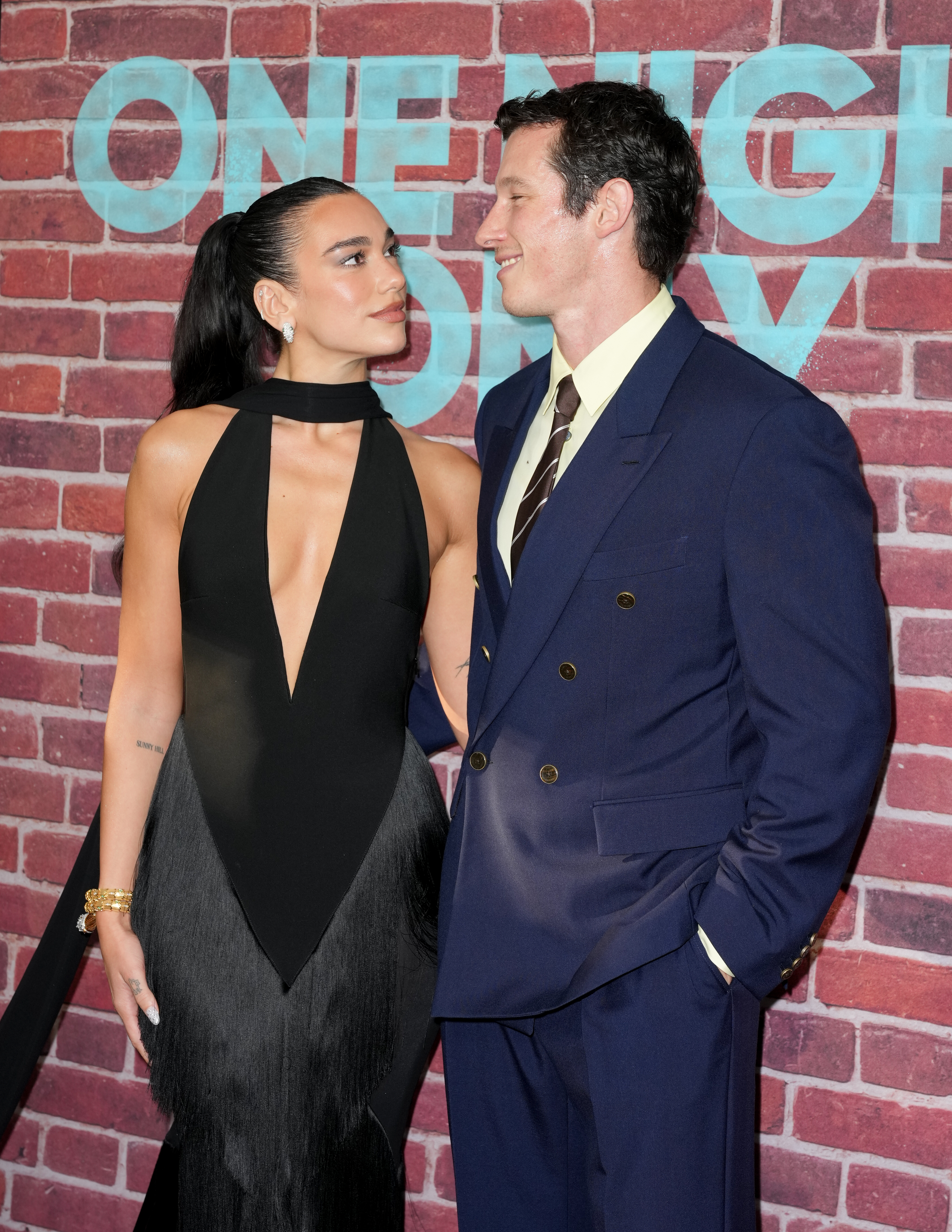A woman in a black halter gown and a man in a navy double-breasted suit smile at each other in front of a brick wall backdrop reading 'One Night Only.'