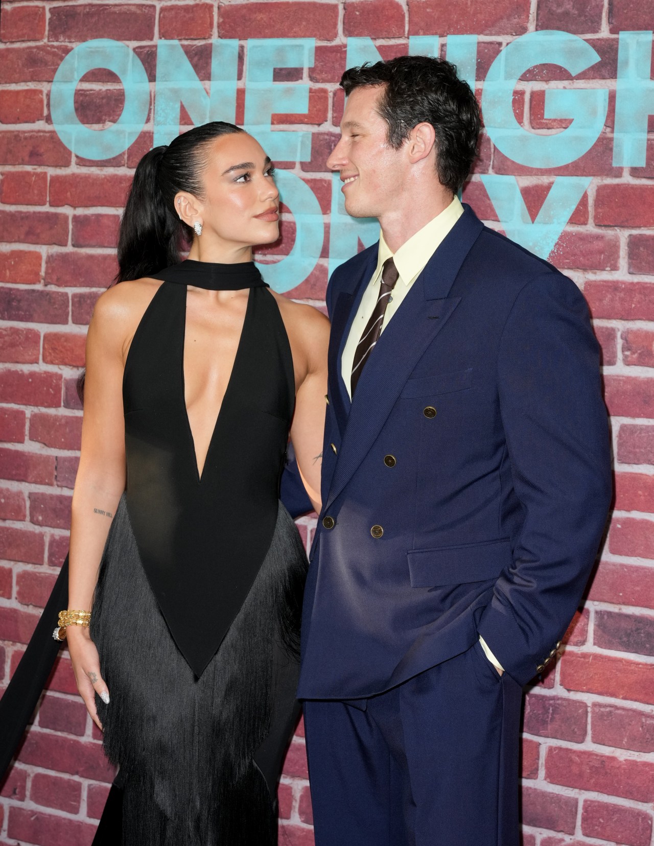 A woman in a black halter gown and a man in a navy double-breasted suit smile at each other in front of a brick wall backdrop reading 'One Night Only.'