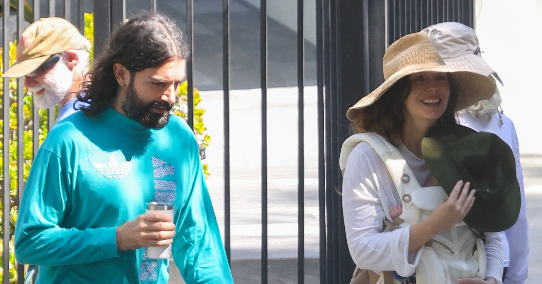 A smiling woman in a wide-brim hat carries a newborn in a front carrier while walking beside a man in a teal long-sleeve shirt holding a tumbler, with a white dog nearby.