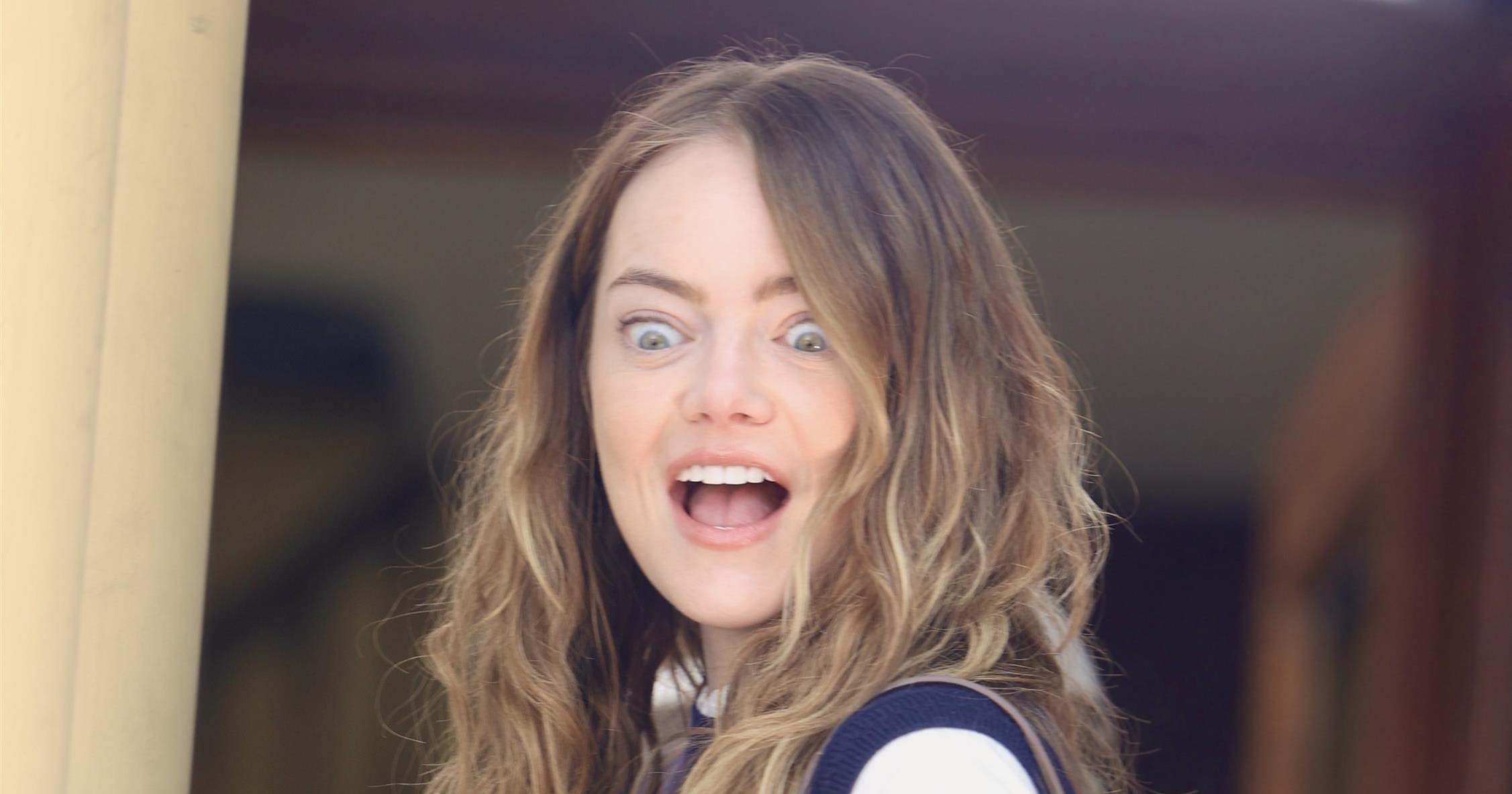 Woman with long wavy brown hair laughing in surprise, wearing a navy vest over a white shirt and carrying a brown suede tote bag.