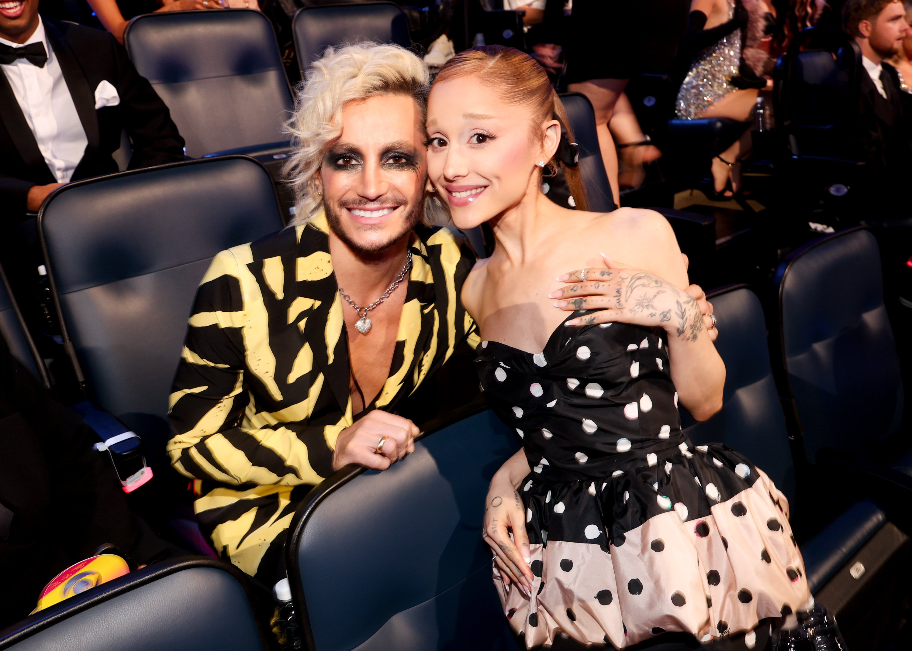 Frankie Grande in a yellow and black blazer and Ariana Grande in a polka-dot dress smile together in the audience at the 2025 MTV VMAs.