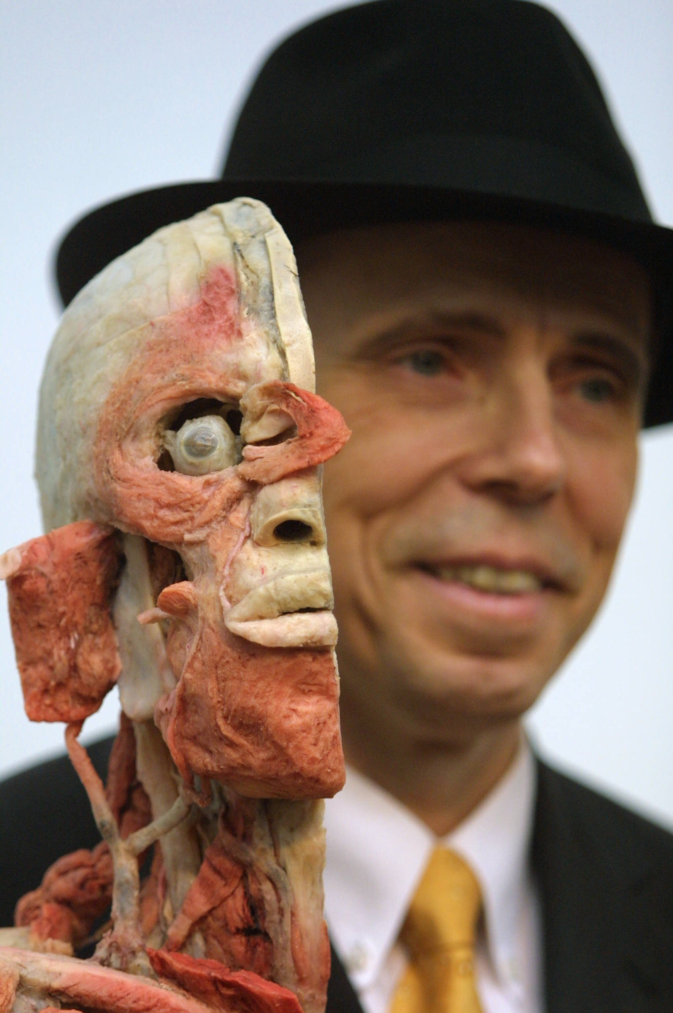Man in a black fedora and suit holding a plastinated dissected human head up close to the camera, smiling in the background.