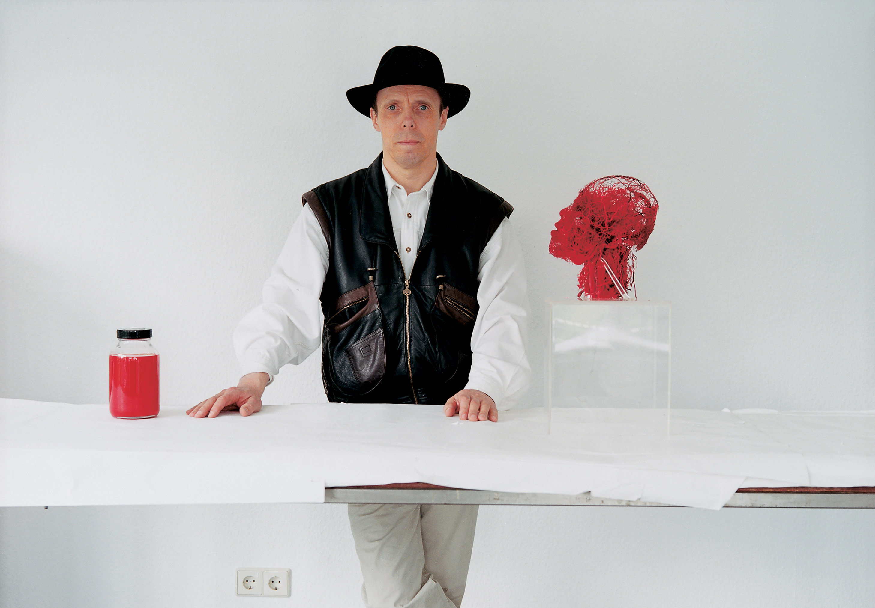 Man in black hat and leather vest stands behind a white table with a red jar and a red mesh human head sculpture on a clear block.