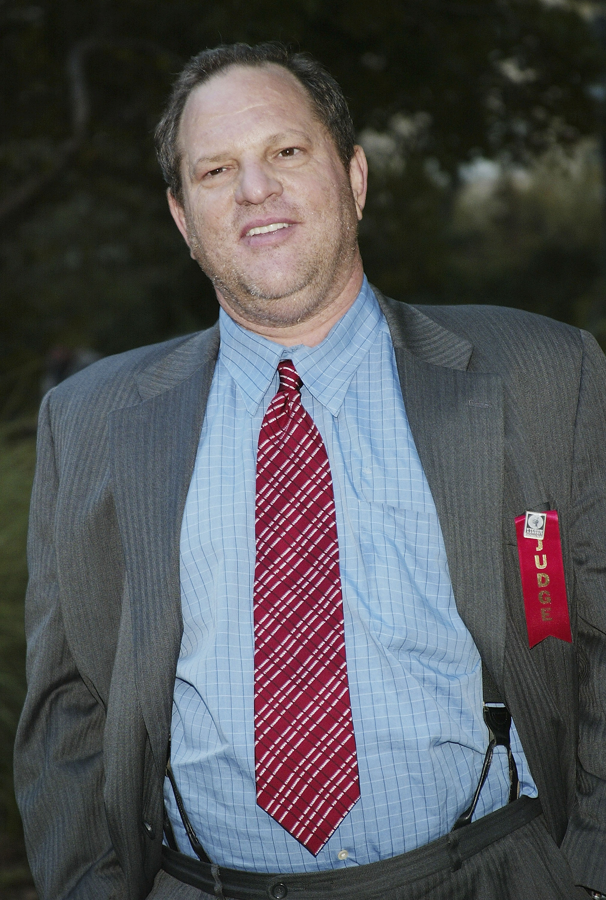 Harvey Weinstein in a grey suit, blue shirt, and red tie with a red Judge ribbon pinned to his lapel, outdoors at an event.