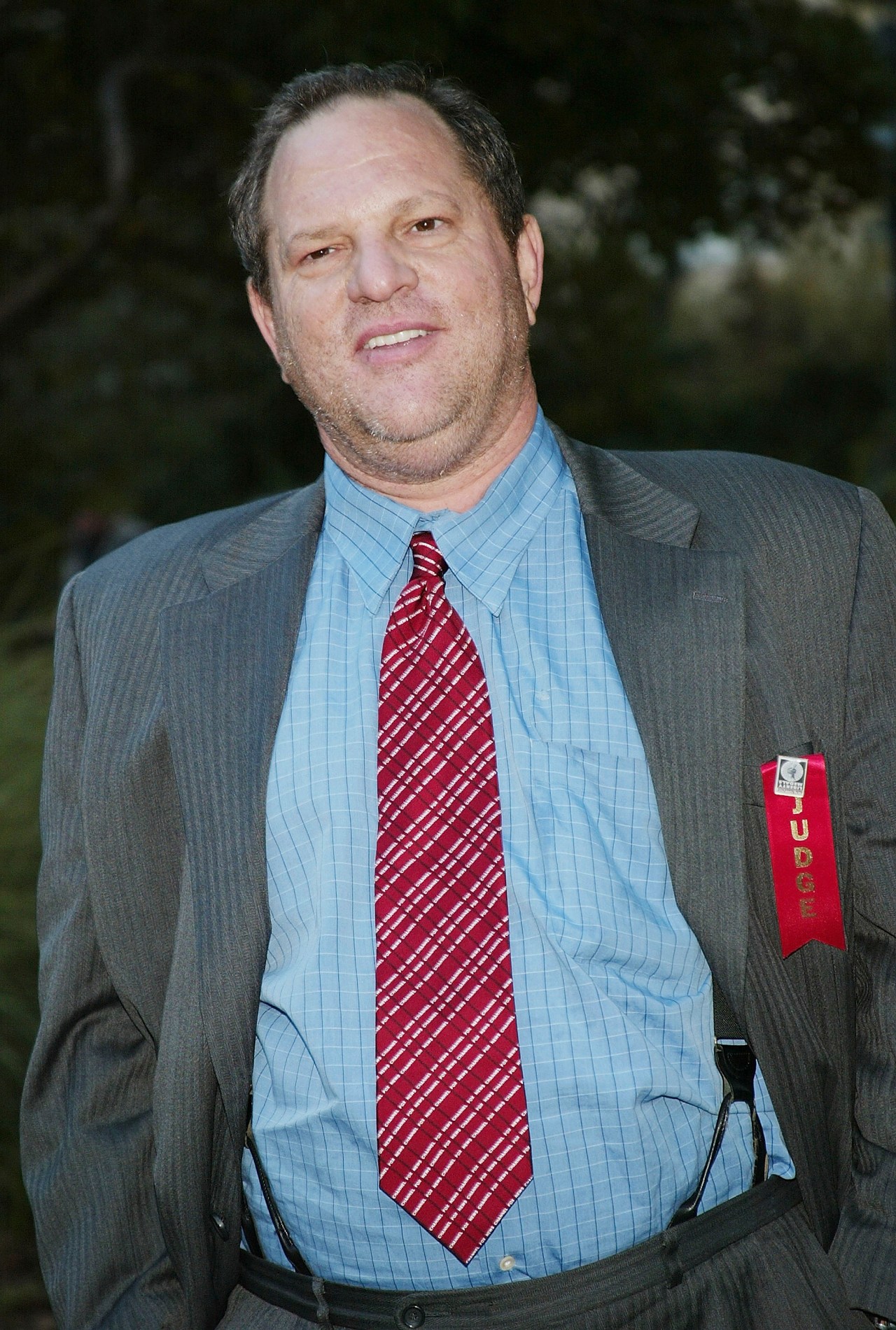 Harvey Weinstein in a grey suit, blue shirt, and red tie with a red Judge ribbon pinned to his lapel, outdoors at an event.