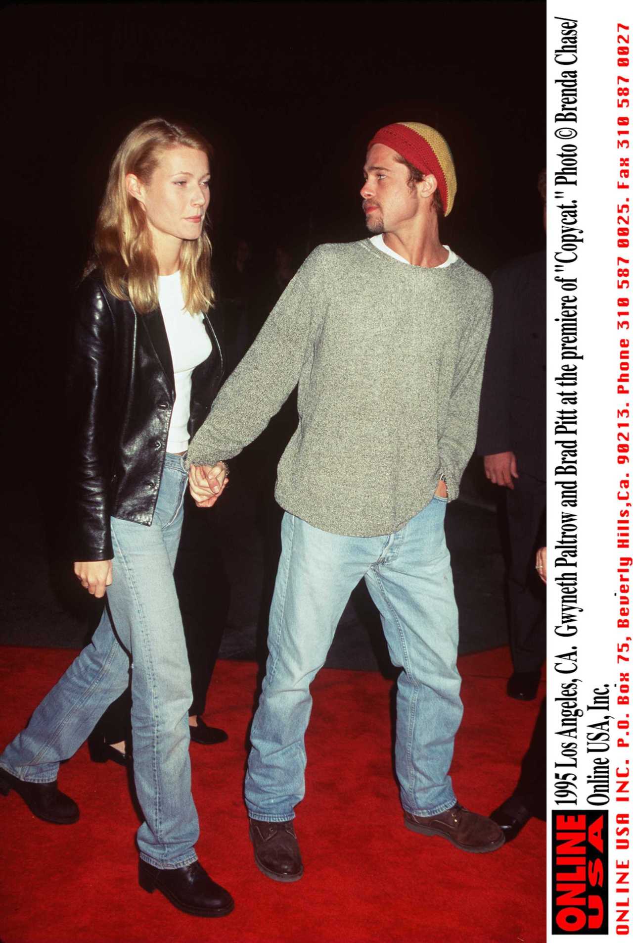 Gwyneth Paltrow and Brad Pitt holding hands on a red carpet, she in a leather jacket and jeans, he in a grey sweater and knit beanie.