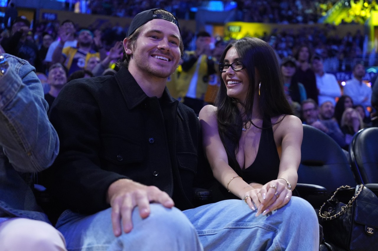 Man in black jacket and backward cap sitting courtside at a basketball game next to a smiling dark-haired woman wearing glasses.