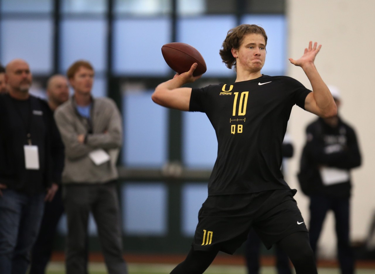 Quarterback wearing Oregon number 10 pro day uniform throwing a football indoors at a scouting event with observers in the background.