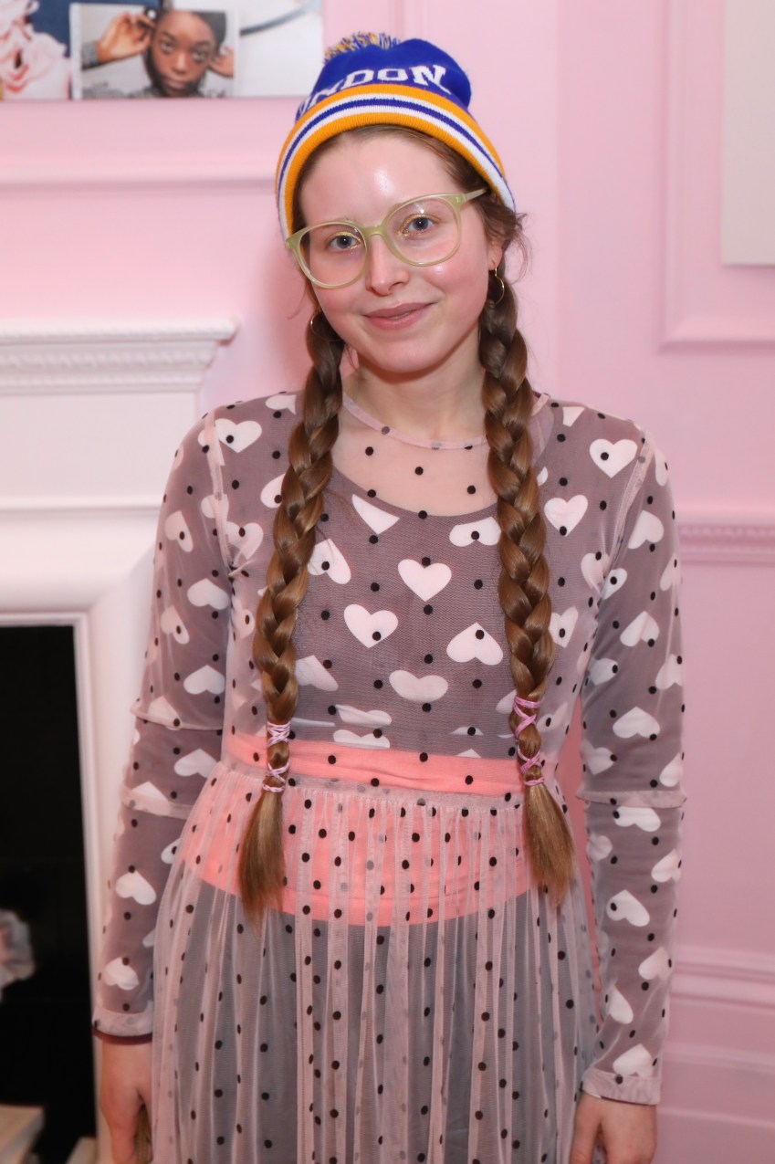 Jessie Cave wearing a blue London beanie, green glasses, twin braids, and a heart-print polka-dot dress against a pink wall.