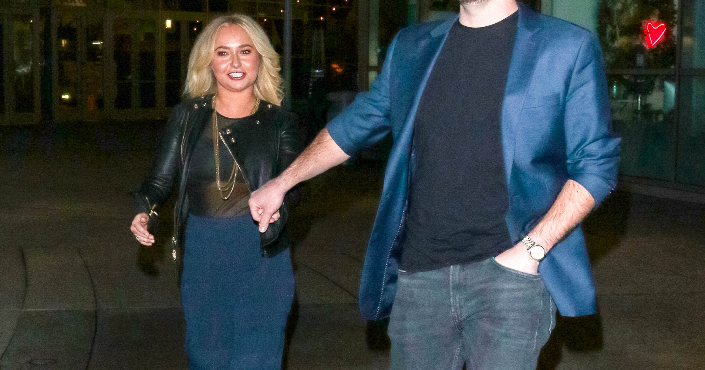 A blonde woman and a tall man in a blue blazer smile while holding hands on a city sidewalk at night.