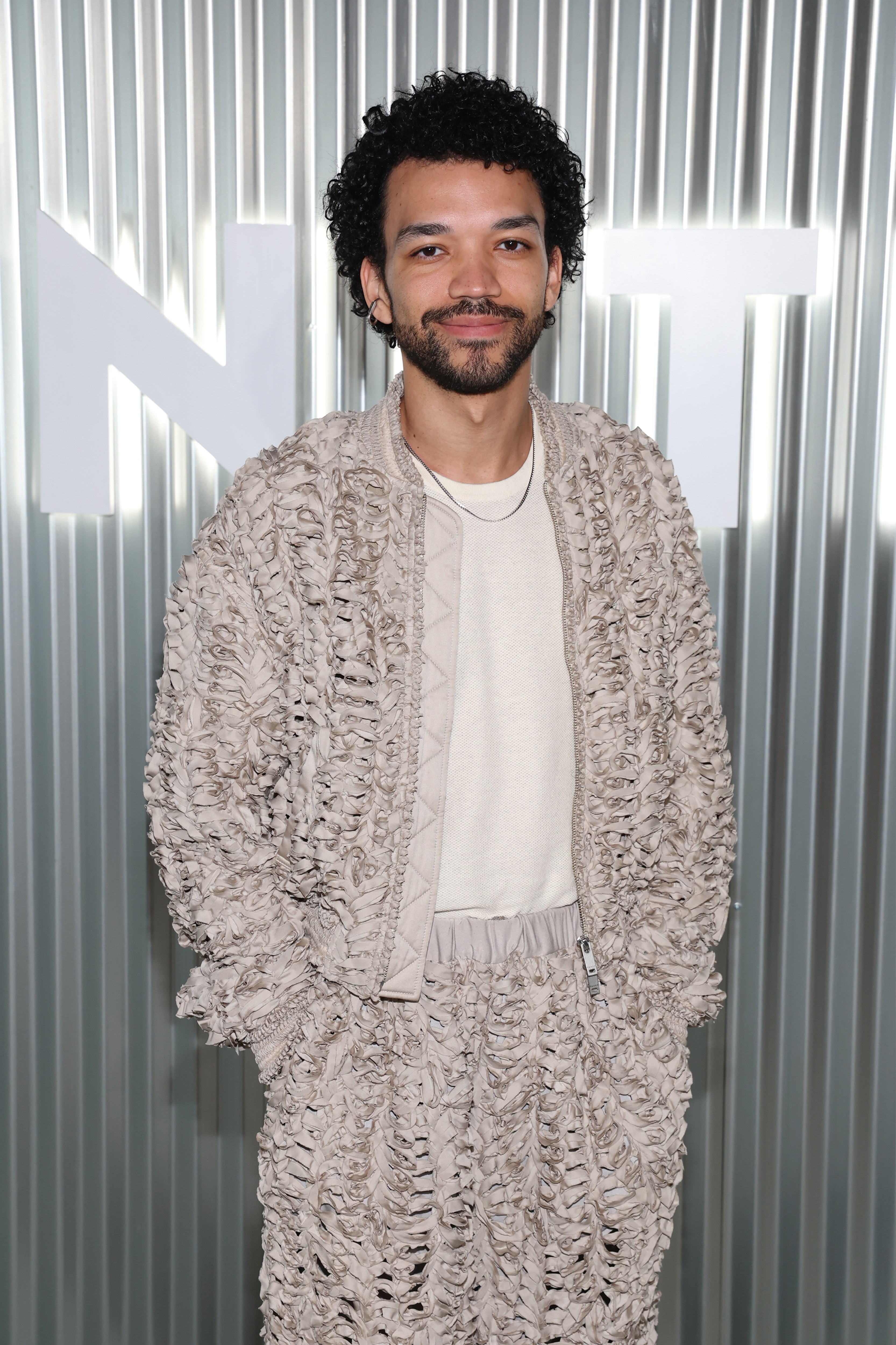 Justice Smith wearing a cream ruffled jacket and matching pants, smiling at a Vanity Fair event in Los Angeles.