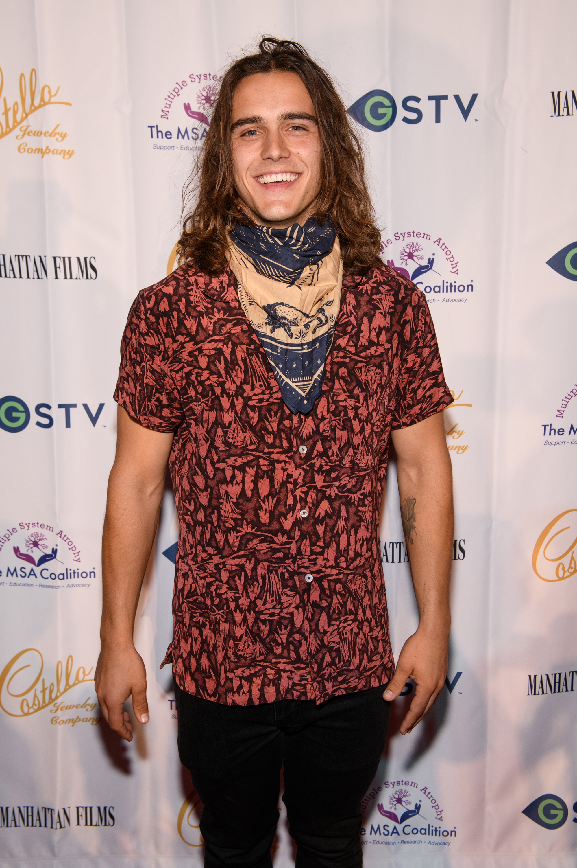 Charlie Gillespie with long brown hair, wearing a dark floral shirt and neck scarf, smiling at a charity event red carpet.