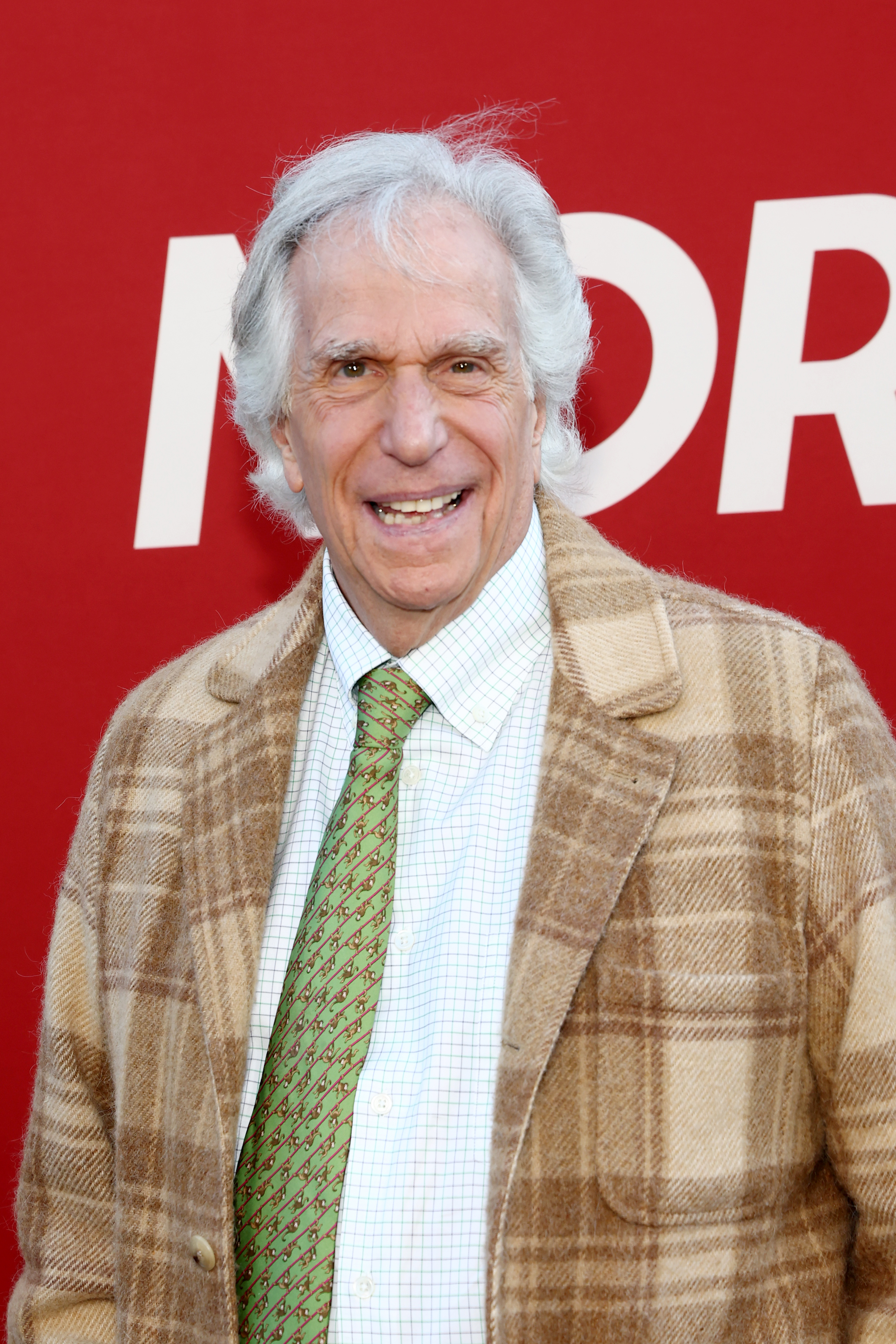 Older man with white hair wearing a tan plaid blazer and green tie smiling in front of a red step-and-repeat backdrop.
