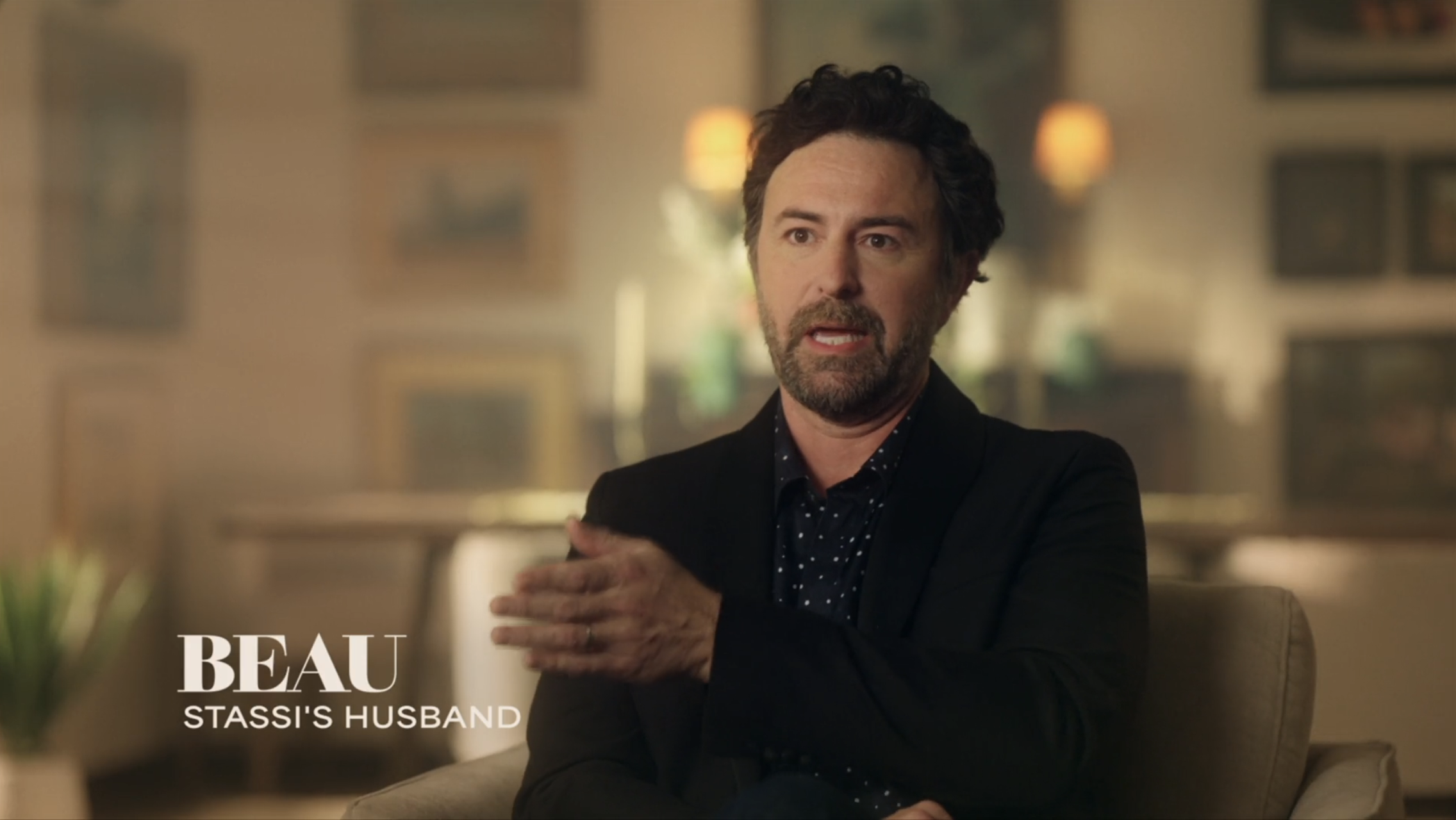 Click to open the image gallery A dark-haired man in a black blazer sits in a chair speaking, with a lower-third graphic reading 'Beau – Stassi's Husband.'