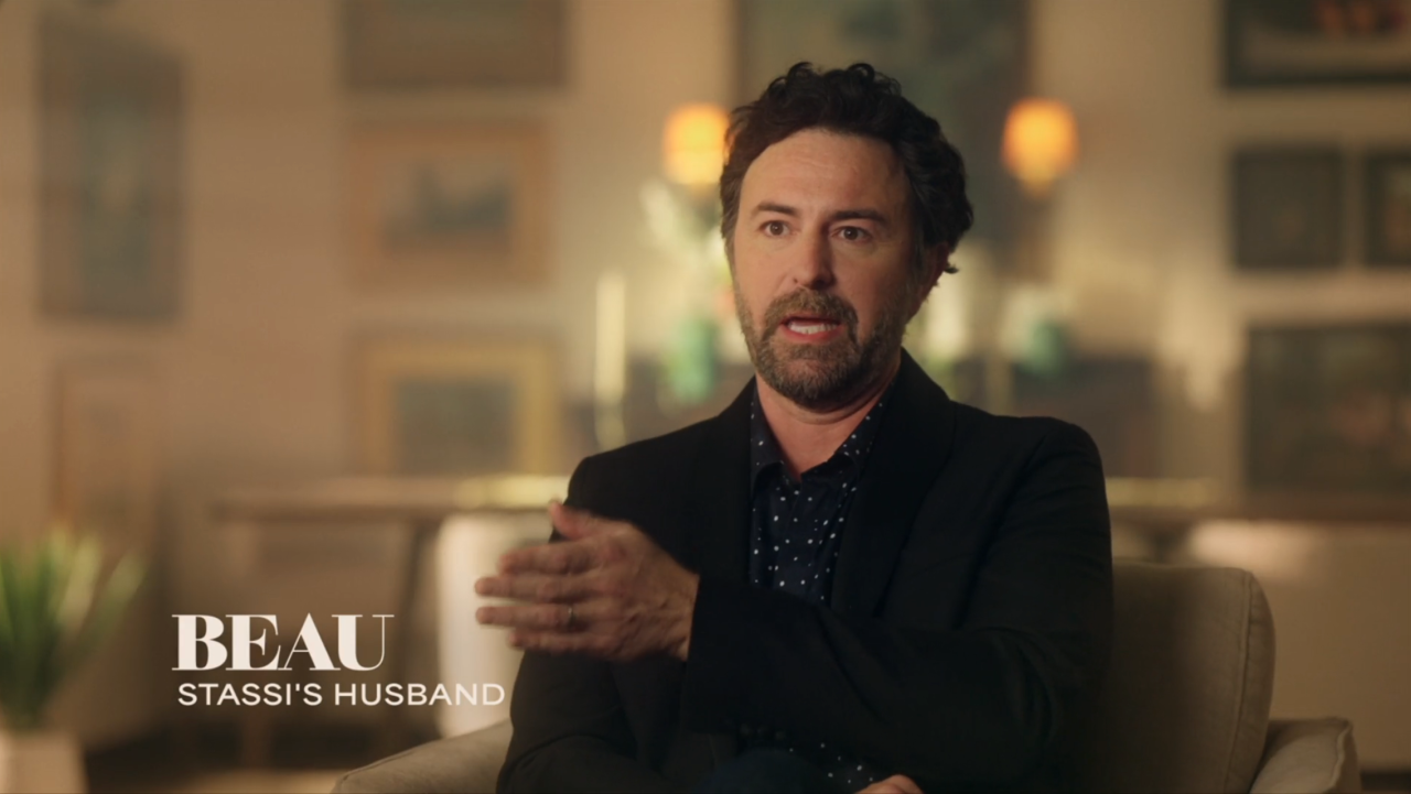 A dark-haired man in a black blazer sits in a chair speaking, with a lower-third graphic reading 'Beau – Stassi's Husband.'