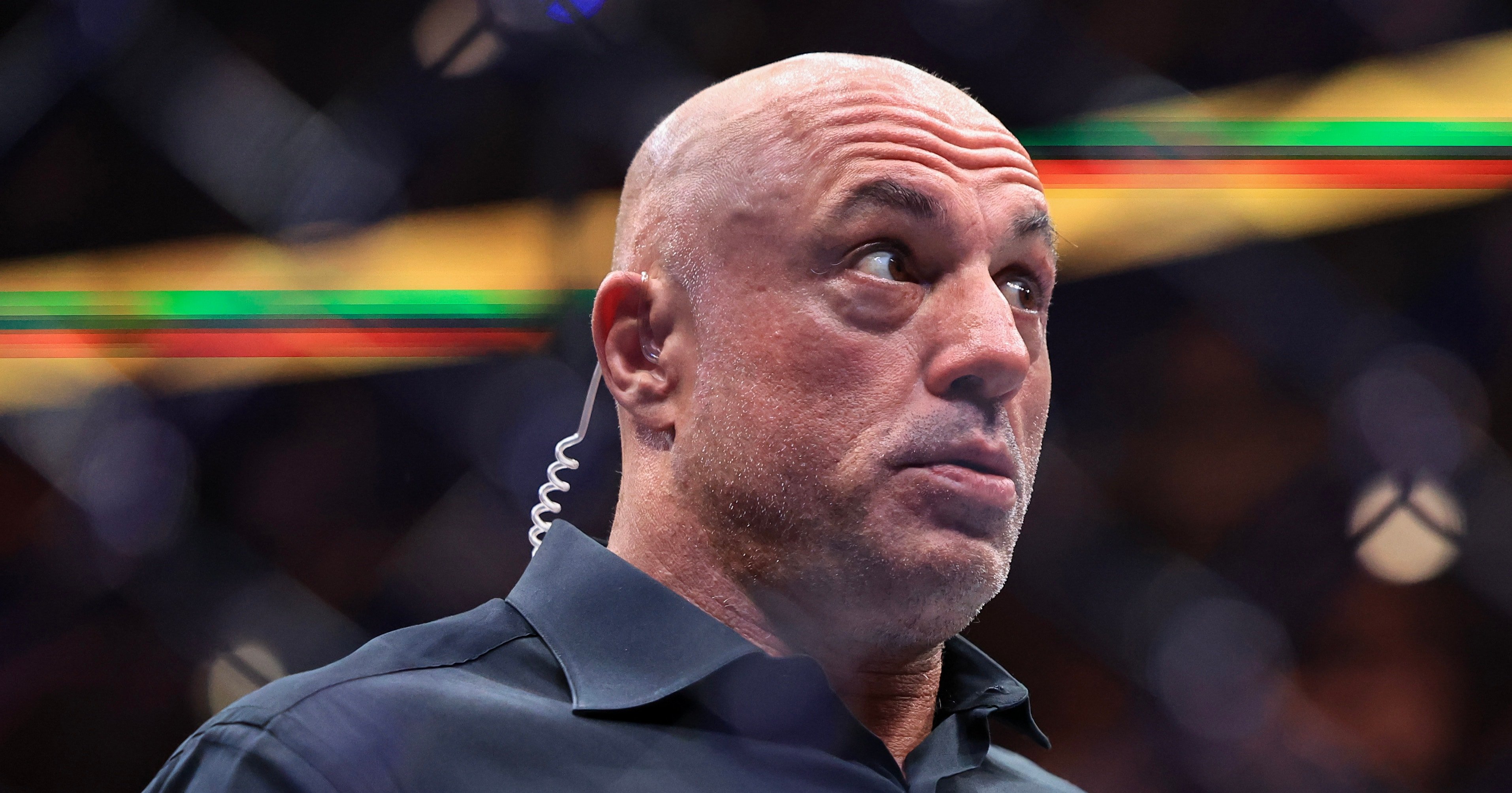 Bald man in a dark shirt with an earpiece cord looks up and to the side with a surprised expression at a UFC event.