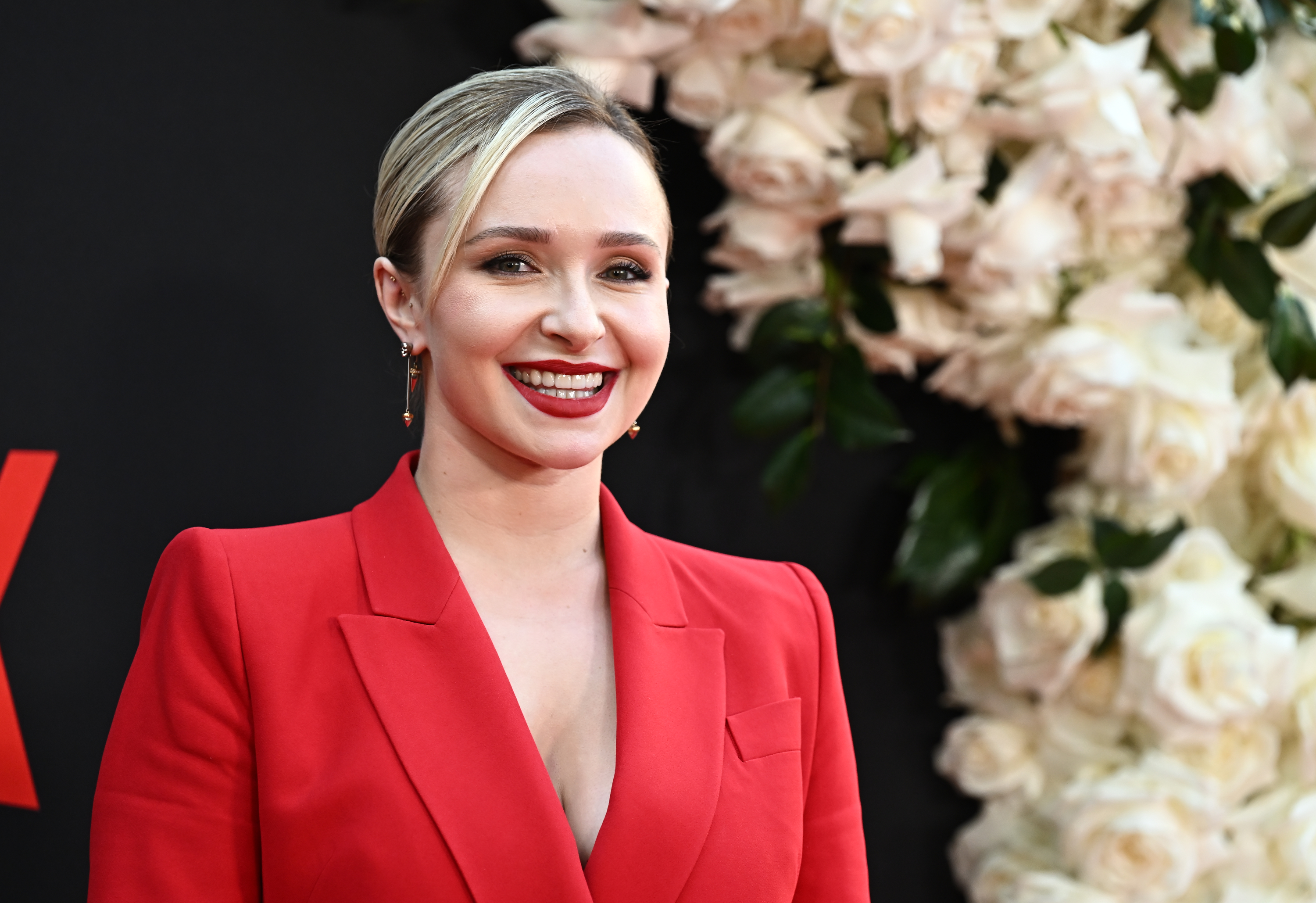Hayden Panettiere smiling in a red blazer at the "Blonde" premiere, with white roses in the background.