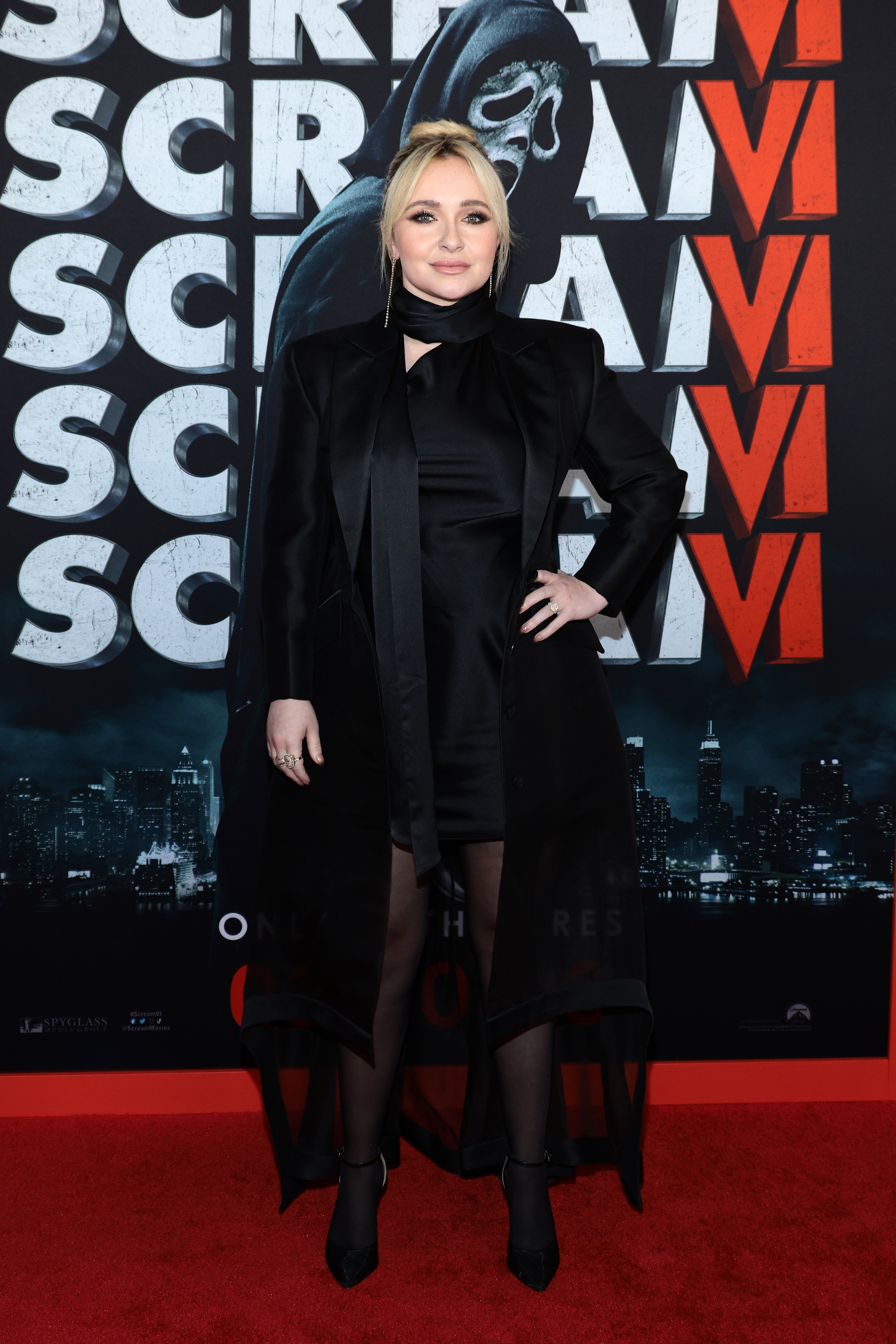 Hayden Panettiere in a black satin dress and coat posing on the red carpet in front of a Scream VI backdrop.