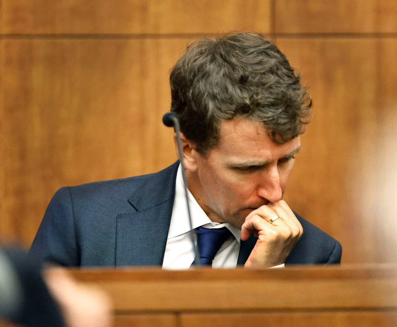 A man in a navy suit and blue tie sits in a courtroom with his hand raised to his mouth, head bowed, in an emotional reaction.