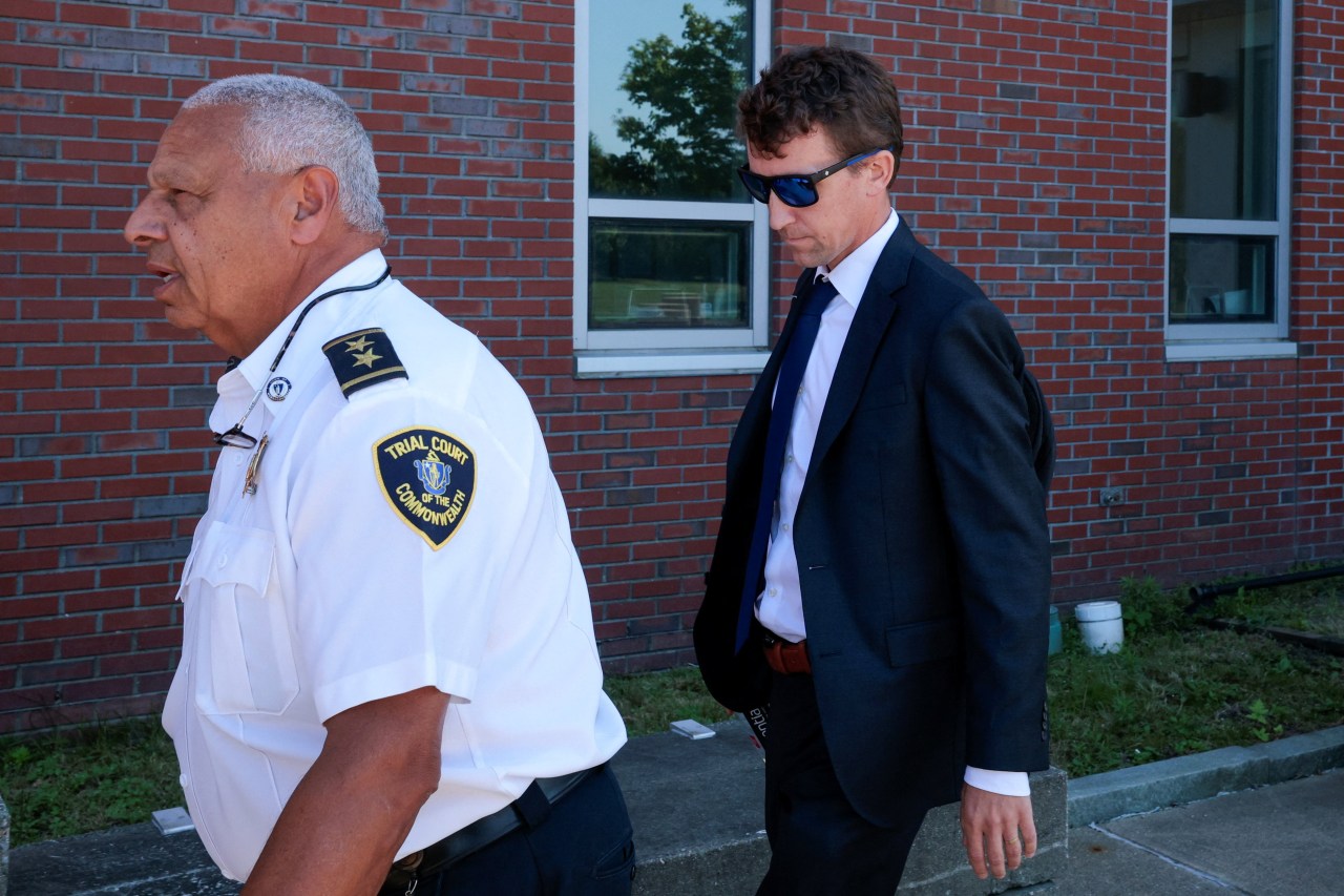 A man in a dark suit and blue tie wearing sunglasses walks alongside a court officer in a white uniform shirt outside a brick courthouse.
