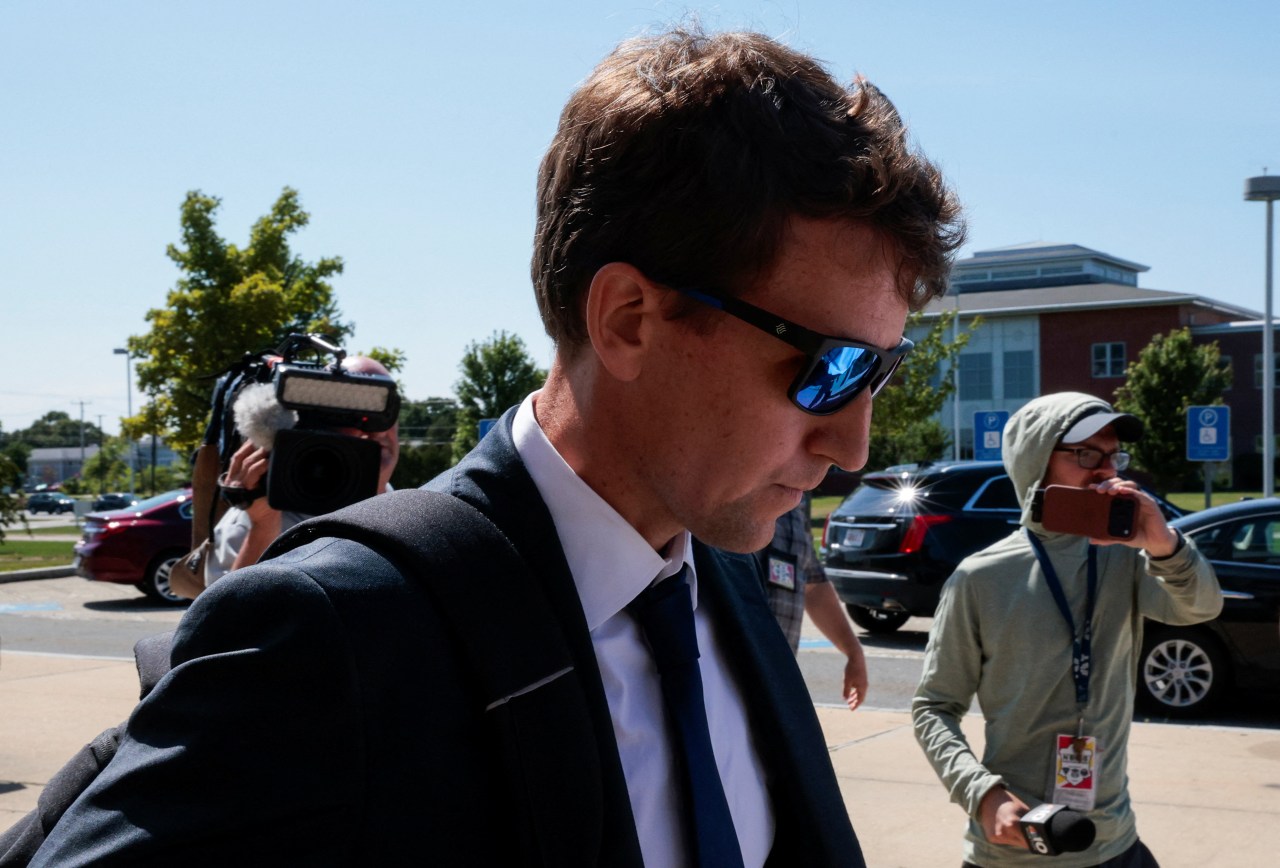 A man in a dark suit and sunglasses walks with his head down through a parking lot as camera crews follow closely behind him.