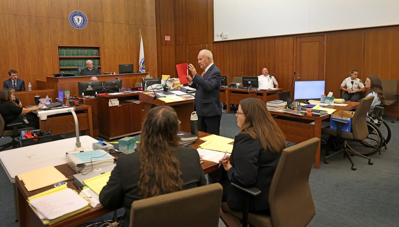 Wide shot of a courtroom showing a man in a navy suit holding a red folder while addressing the court, with attorneys seated at tables and a judge on the bench.