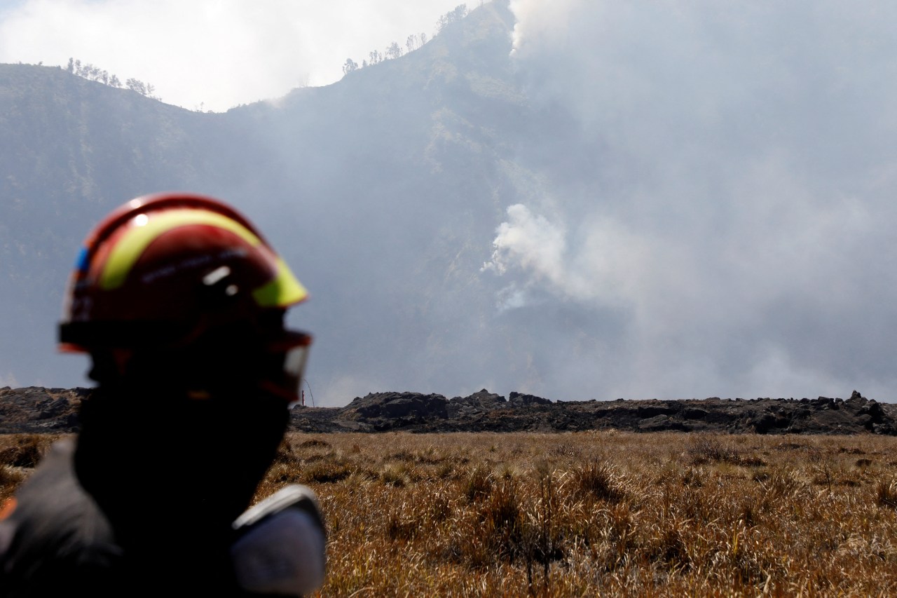 A firefighter in a red and yellow helmet stands in the foreground, looking toward a smoke-covered mountain slope and dry grass plain.