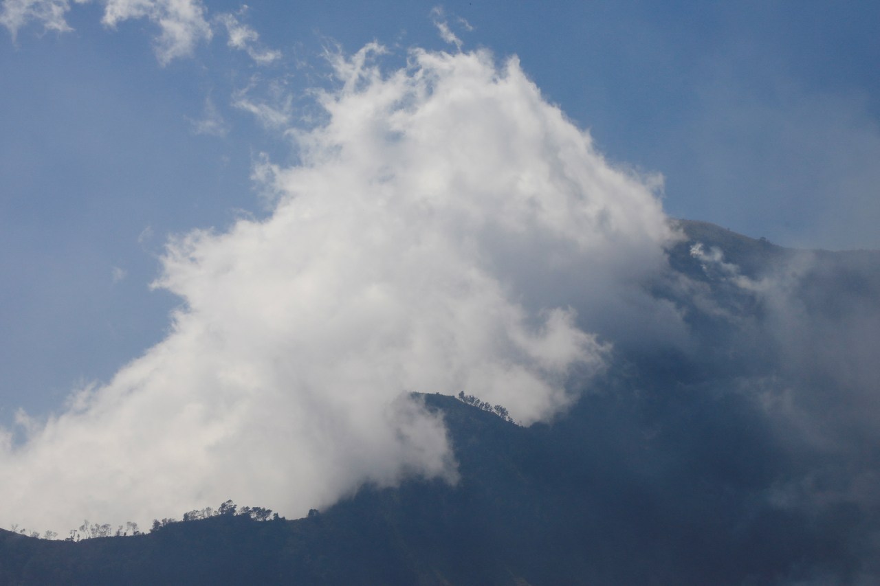 A large white smoke cloud billows over a dark mountain silhouette with scattered trees against a blue sky.