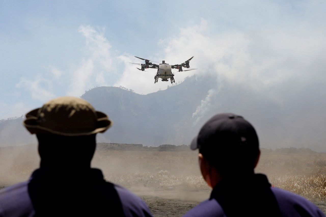 Two people seen from behind watch a large multi-rotor drone hovering in smoky air above a dry plain, with a smoking mountain in the background.