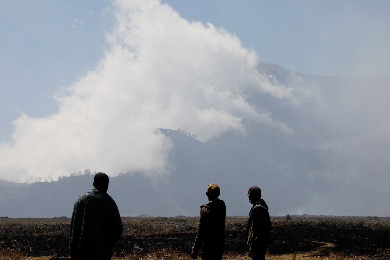 Three people stand in silhouette on a burnt field, looking toward a large smoke cloud rising above a mountain.