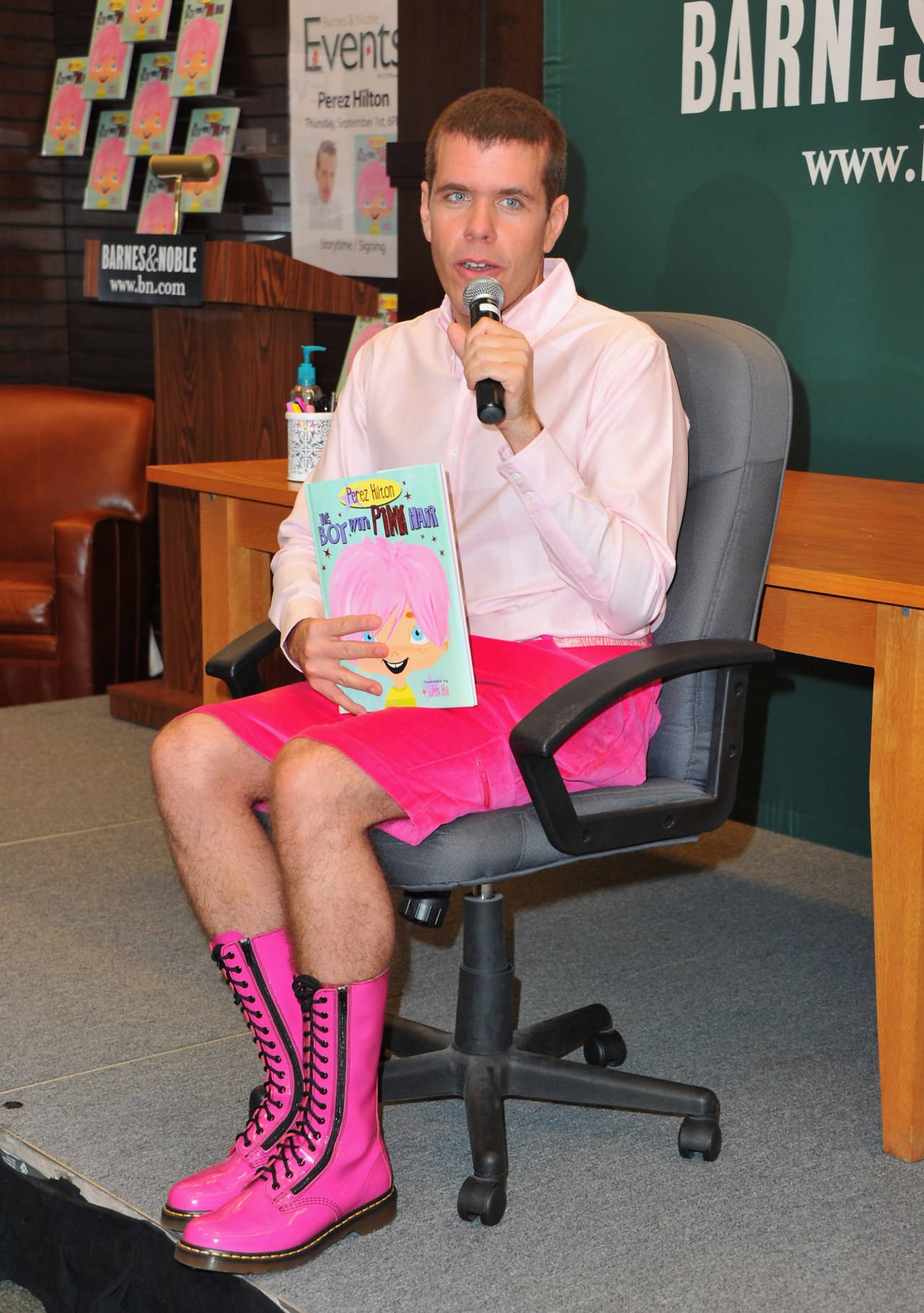 Perez Hilton sitting in an office chair at a Barnes & Noble, holding a microphone and his children's book "The Boy With Pink Hair," wearing pink shorts and tall pink boots.
