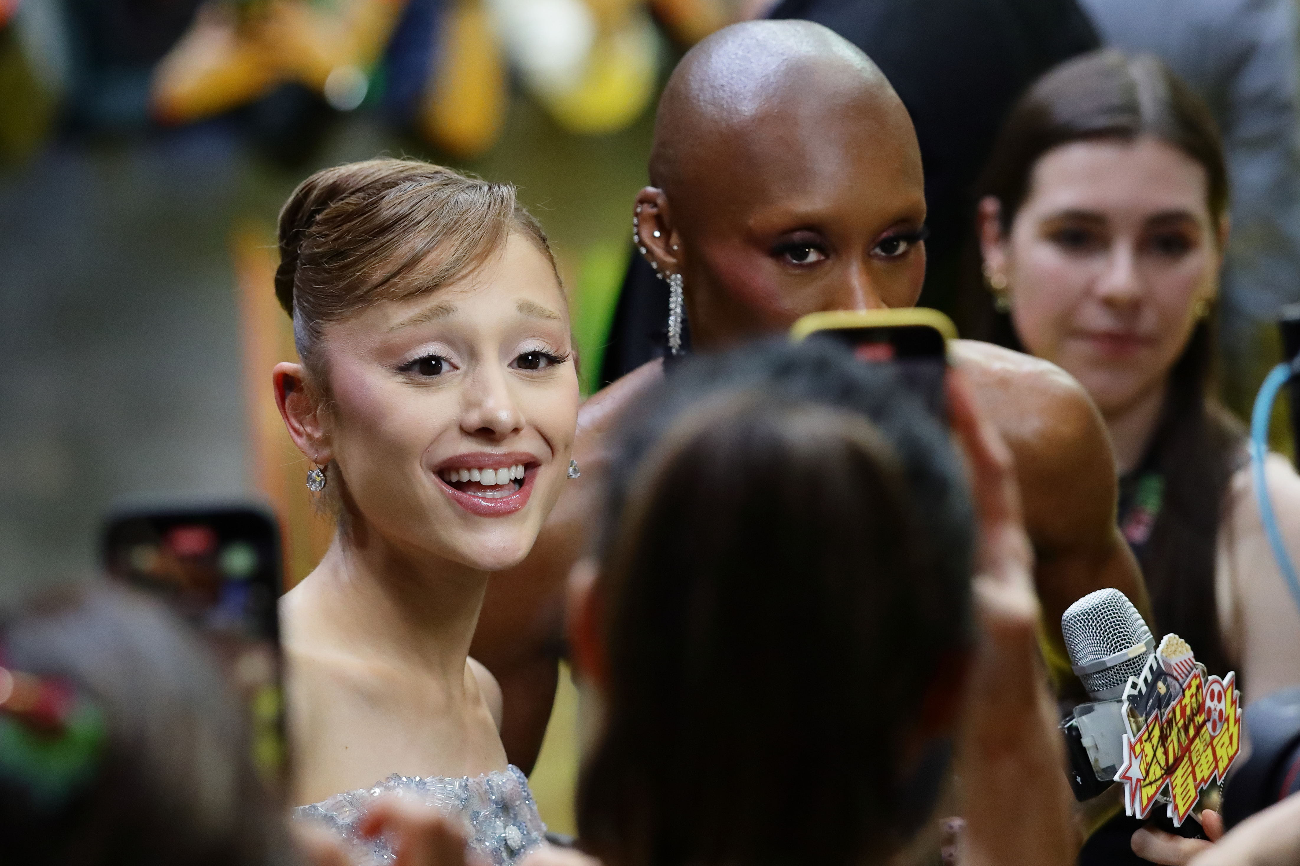 Ariana Grande smiling and speaking into a microphone at a red carpet event, with Cynthia Erivo visible behind her.