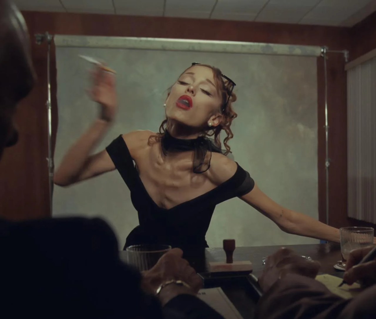 A woman in a black off-shoulder dress and choker with red lips leans back dramatically at a desk in a moody, dimly lit room.