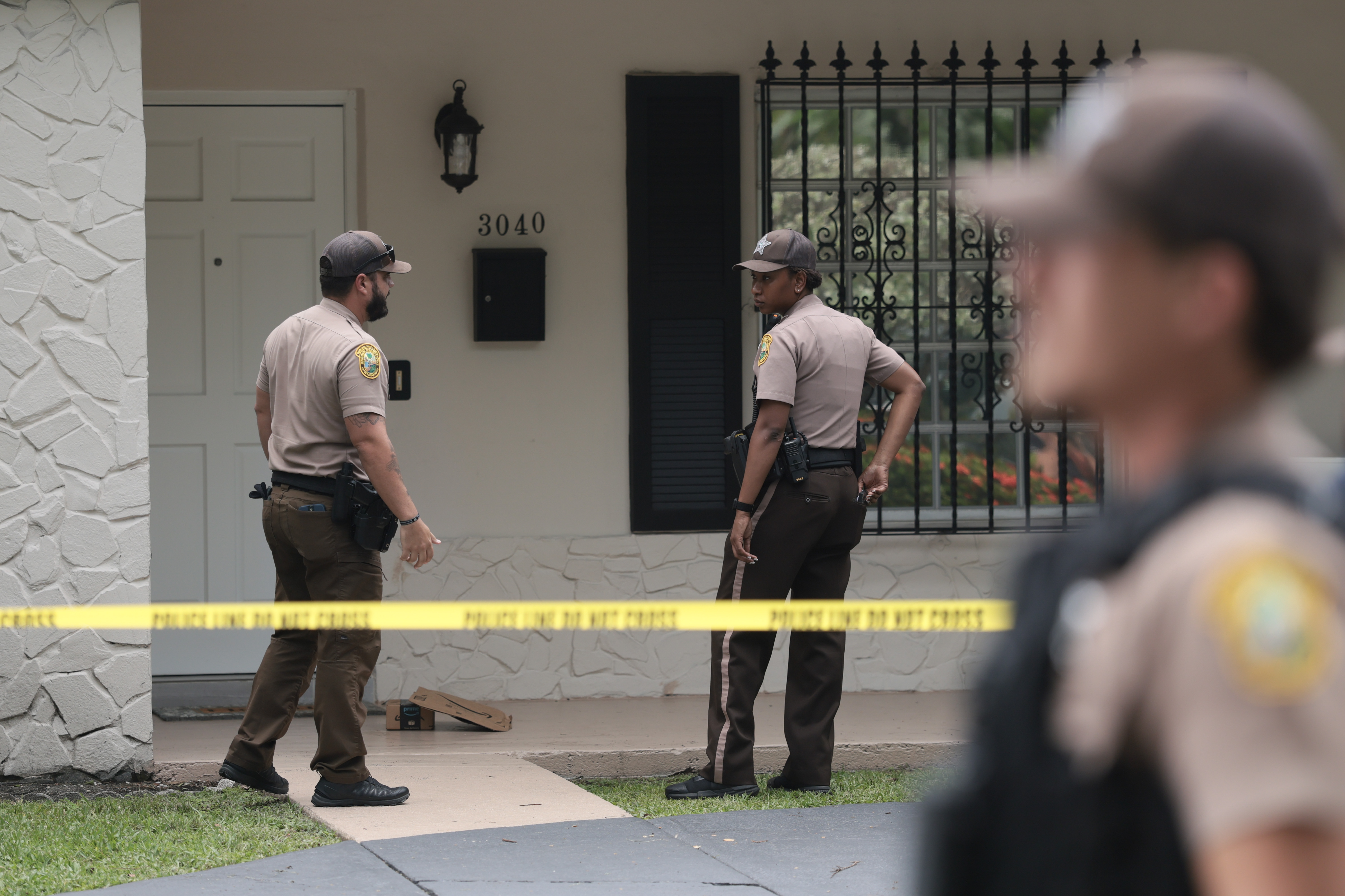 Two Miami-Dade County Sheriff's deputies confer outside a home at address 3040, with yellow police tape strung across the front yard.