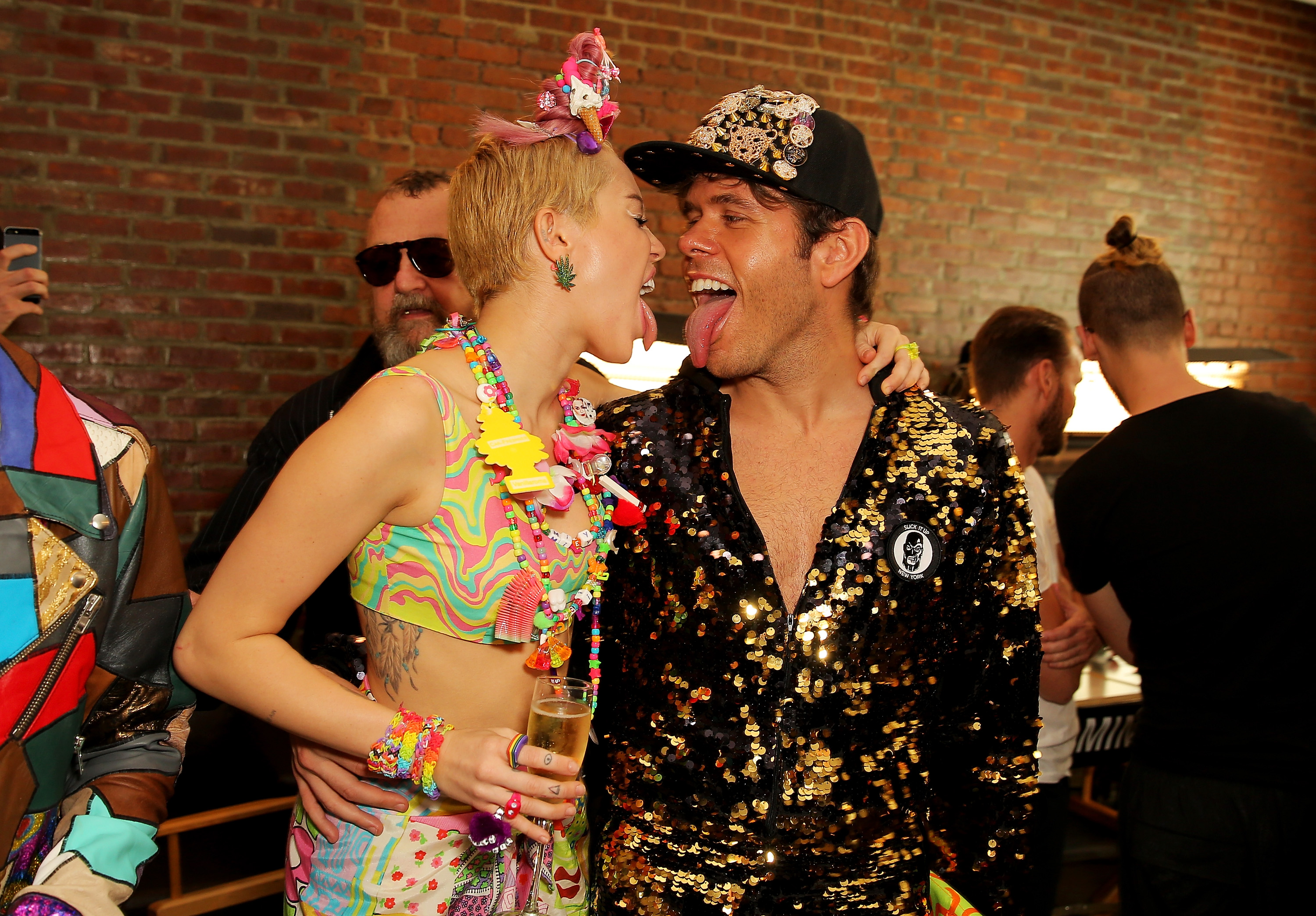 Miley Cyrus in a colorful crop top and candy accessories touches tongues with Perez Hilton in a gold sequin jacket backstage at a fashion show.