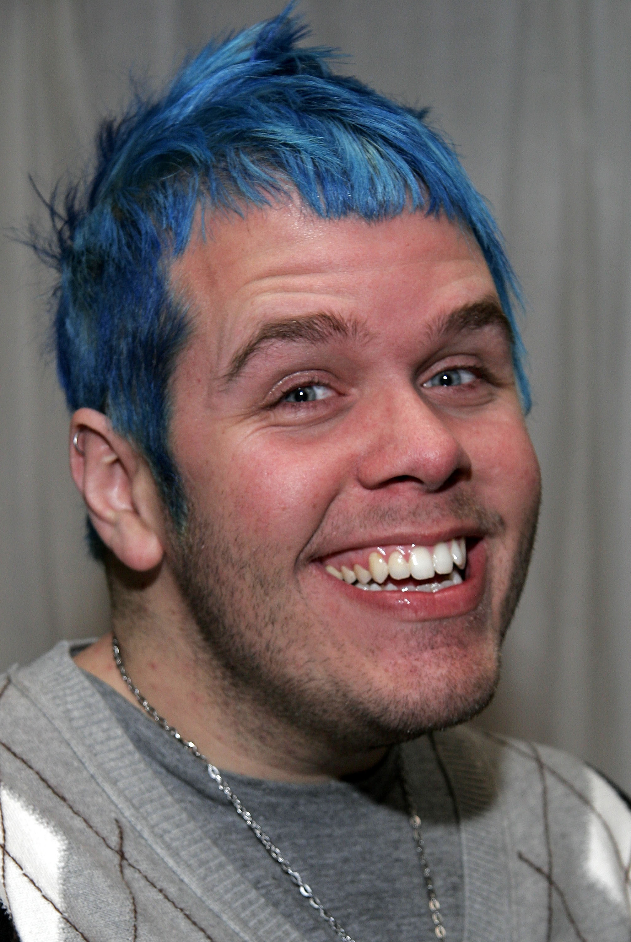 Man with bright blue spiky hair and a silver chain necklace smiling broadly against a gray curtain backdrop.