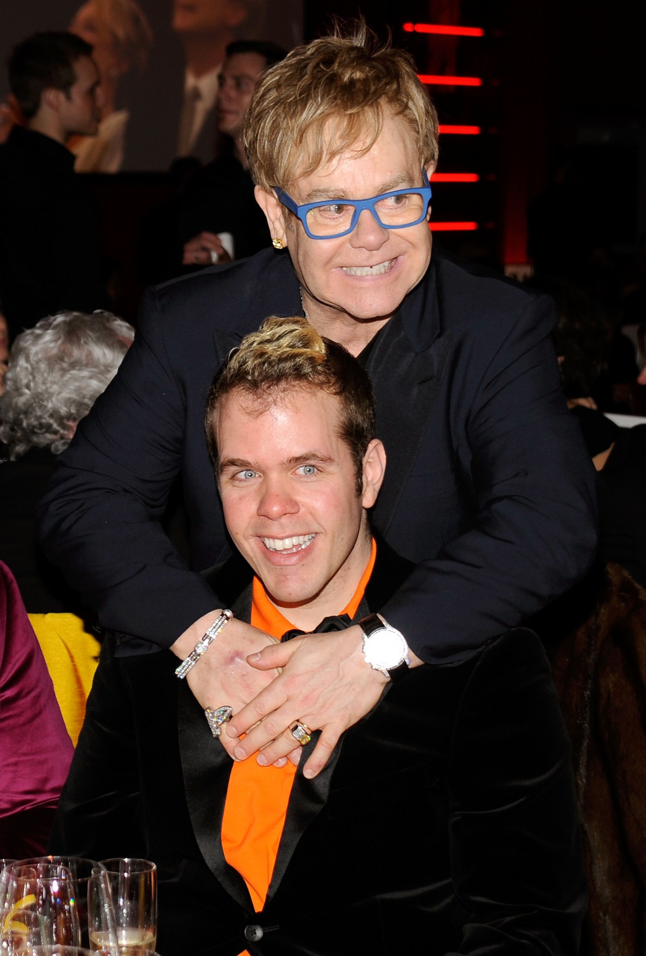 A man in a black blazer and orange tie seated while another man in blue glasses hugs him from behind at an indoor gala event.