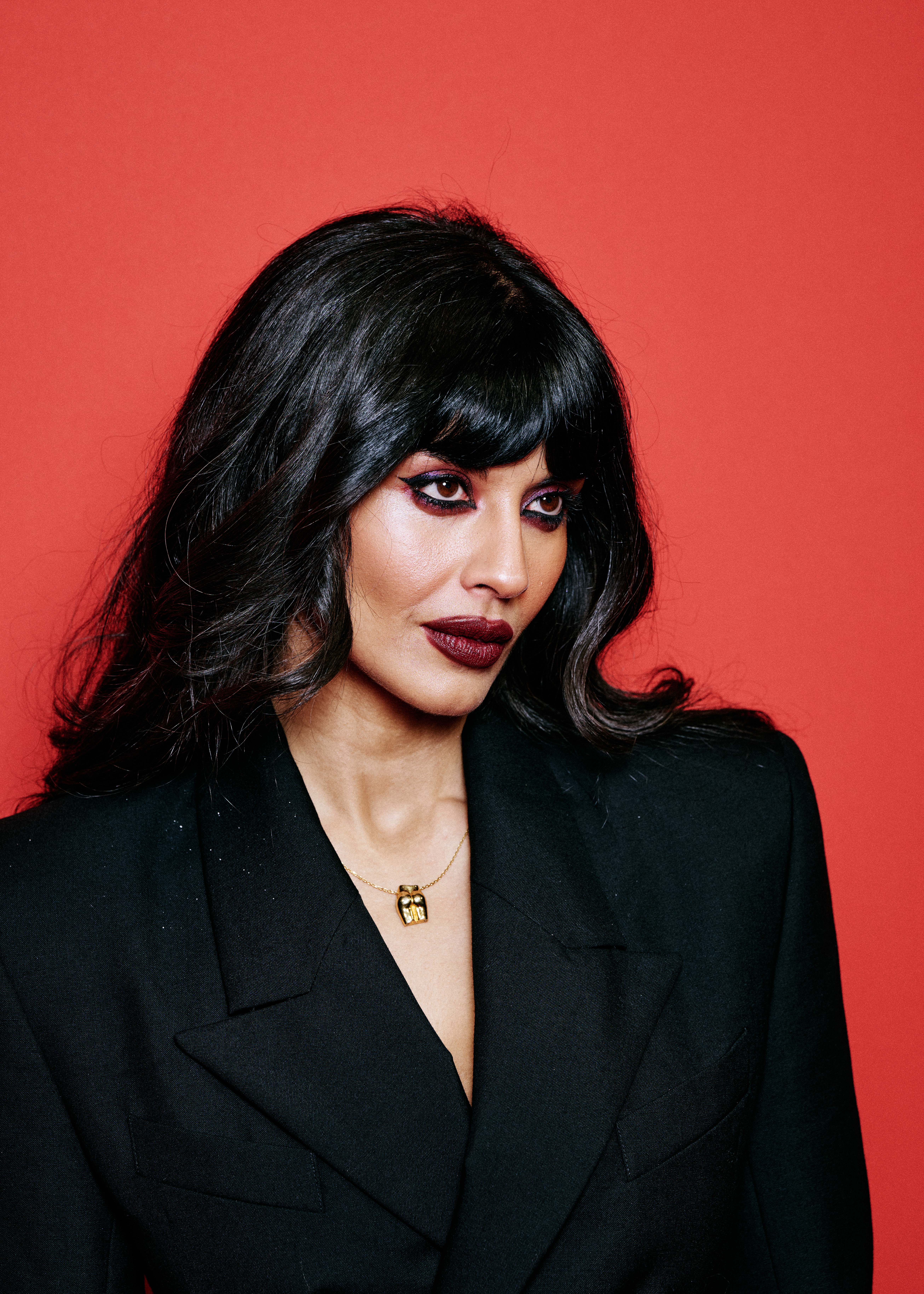 Click to open the image gallery Jameela Jamil in a black blazer and gold necklace with dramatic dark makeup against a red backdrop at the GQ Men of the Year Awards 2025.