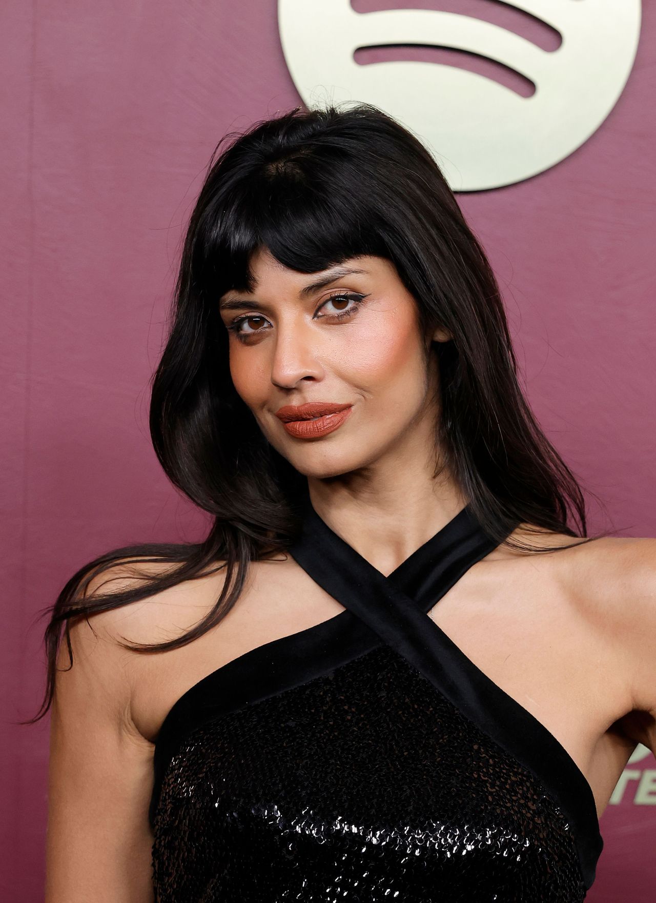 Jameela Jamil with long dark hair and bangs, wearing a black cross-halter top, posing in front of a Spotify logo backdrop