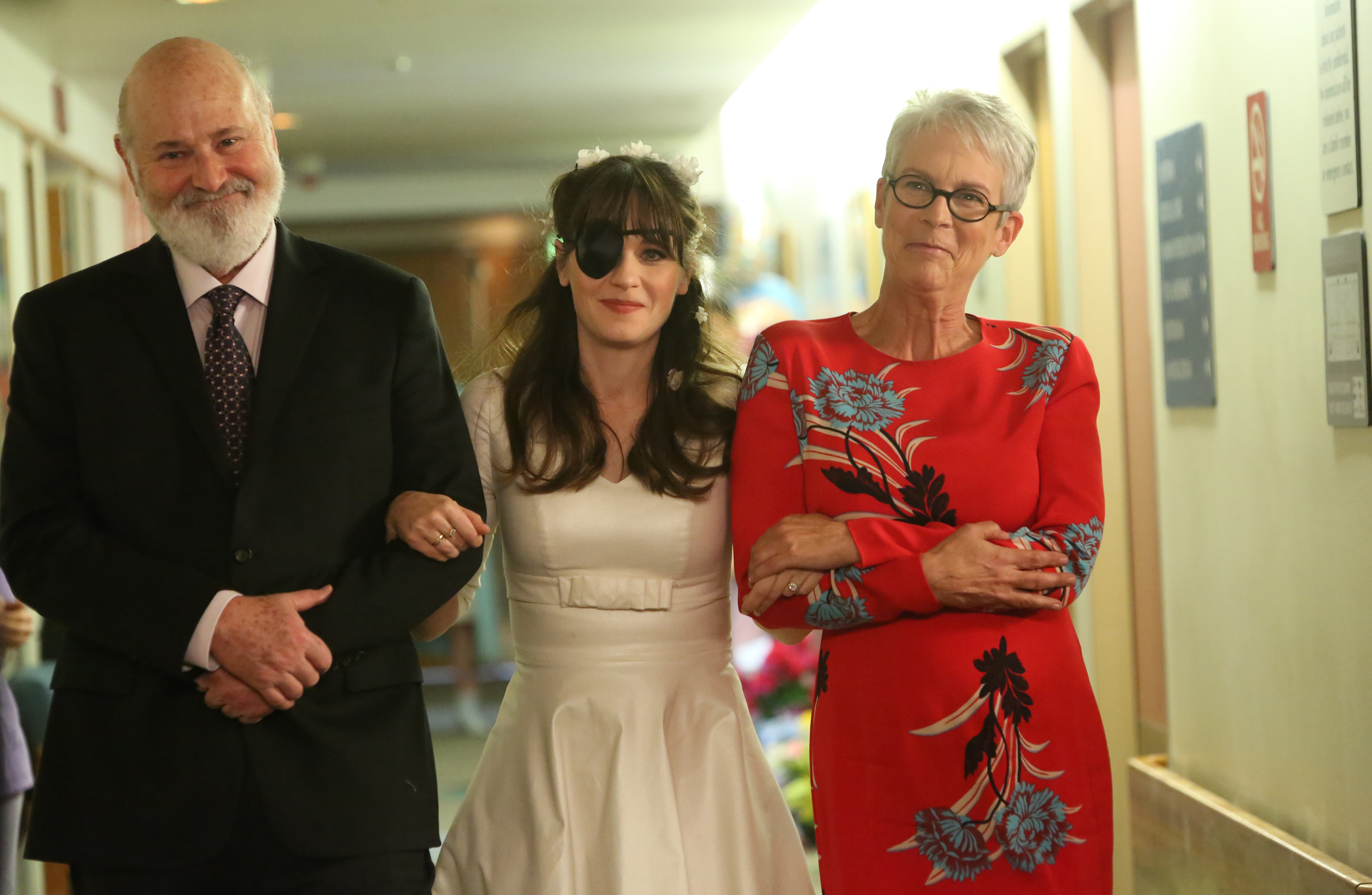 Click to open the image gallery Close-up of Rob Reiner smiling, Zooey Deschanel in a white dress and black eye patch, and Jamie Lee Curtis in a red floral dress with arms crossed, in a hallway.