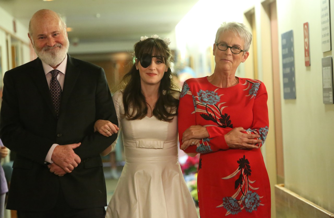 Close-up of Rob Reiner smiling, Zooey Deschanel in a white dress and black eye patch, and Jamie Lee Curtis in a red floral dress with arms crossed, in a hallway.