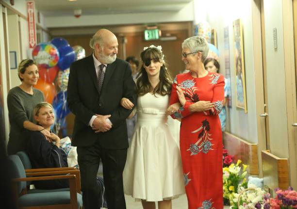 Rob Reiner, Zooey Deschanel in a white wedding dress and eye patch, and Jamie Lee Curtis in a red floral dress walking a hospital hallway.